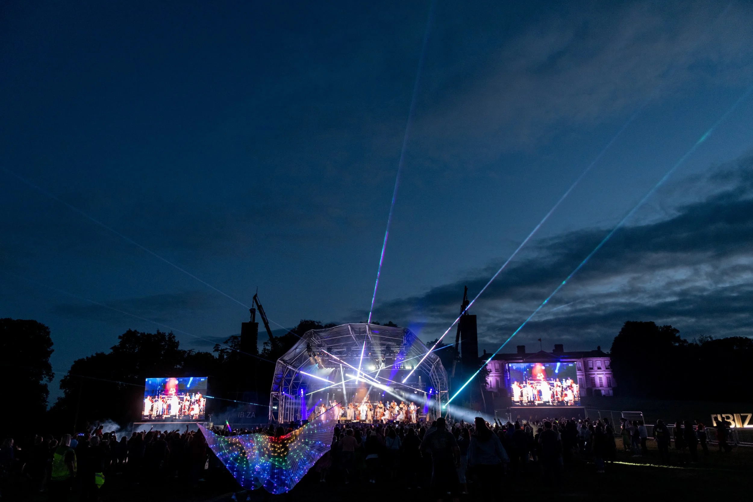 Outdoor concert Ibiza Classic at night with stage, bright lights, large screens, and audience, under a cloudy sky with laser light beams.