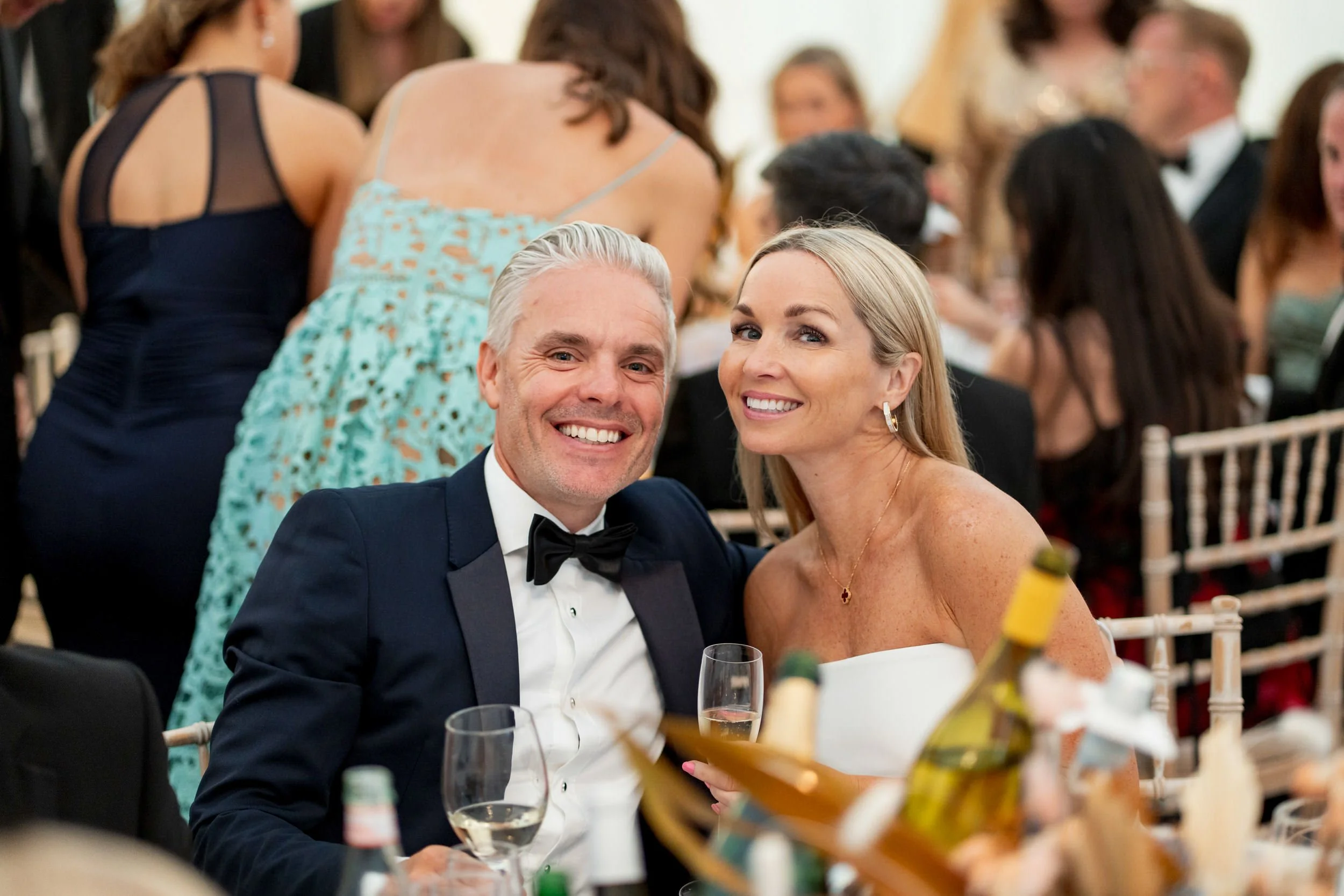 A smiling couple dressed in elegant attire, sitting at a formal event. The man is wearing a tuxedo with a bow tie, and the woman is in a strapless dress with gold earrings, holding wine glasses. The background shows other guests and tables, indicatin