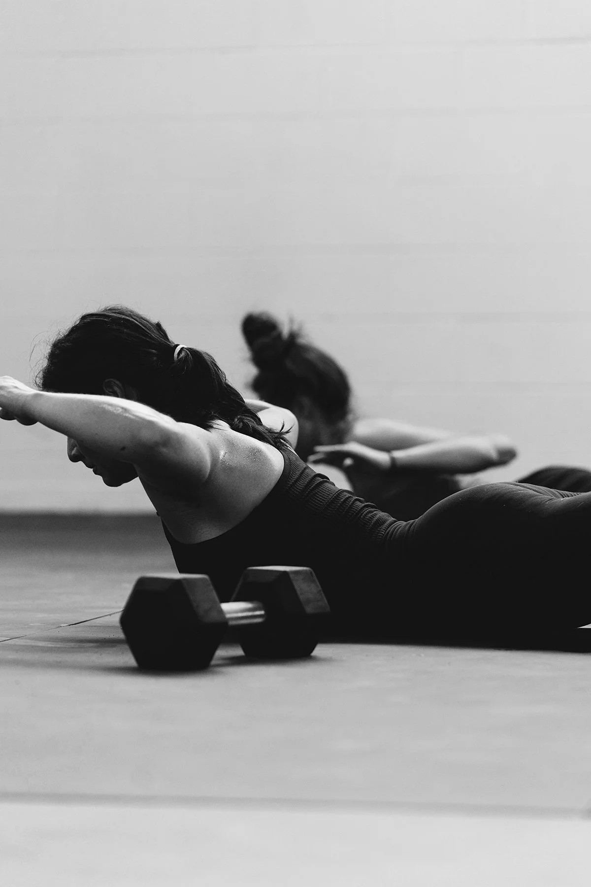 Zwei Frauen beim Yoga-Workout im Fitnessstudio, liegend auf der Matte, während des Bein- und Oberkörpertrainings mit Hanteln, in schwarz-weißer Aufnahme.