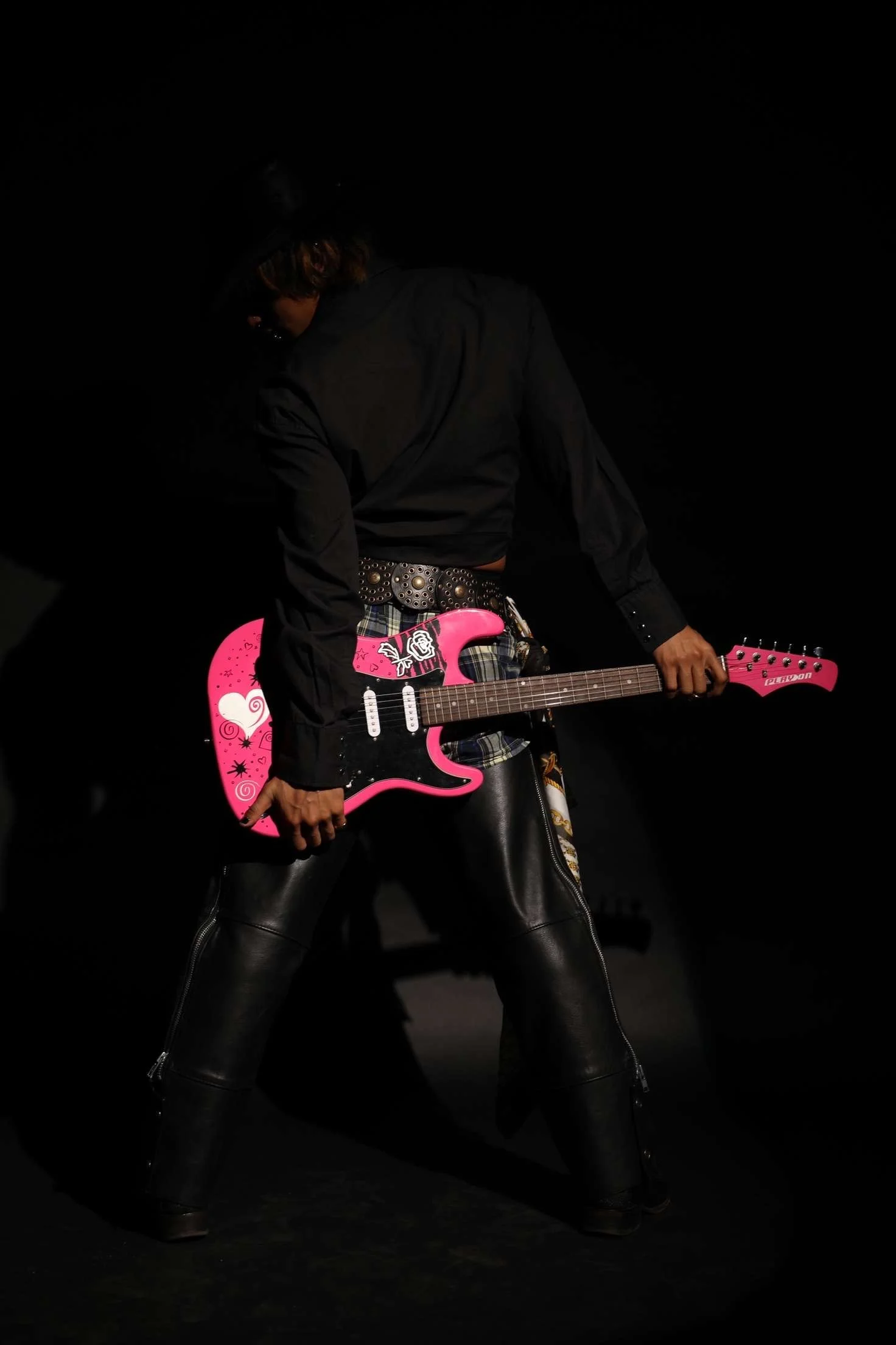 Person dressed in black with leather pants and a plaid skirt, holding a pink electric guitar with heart and swirl designs, on a dark stage.