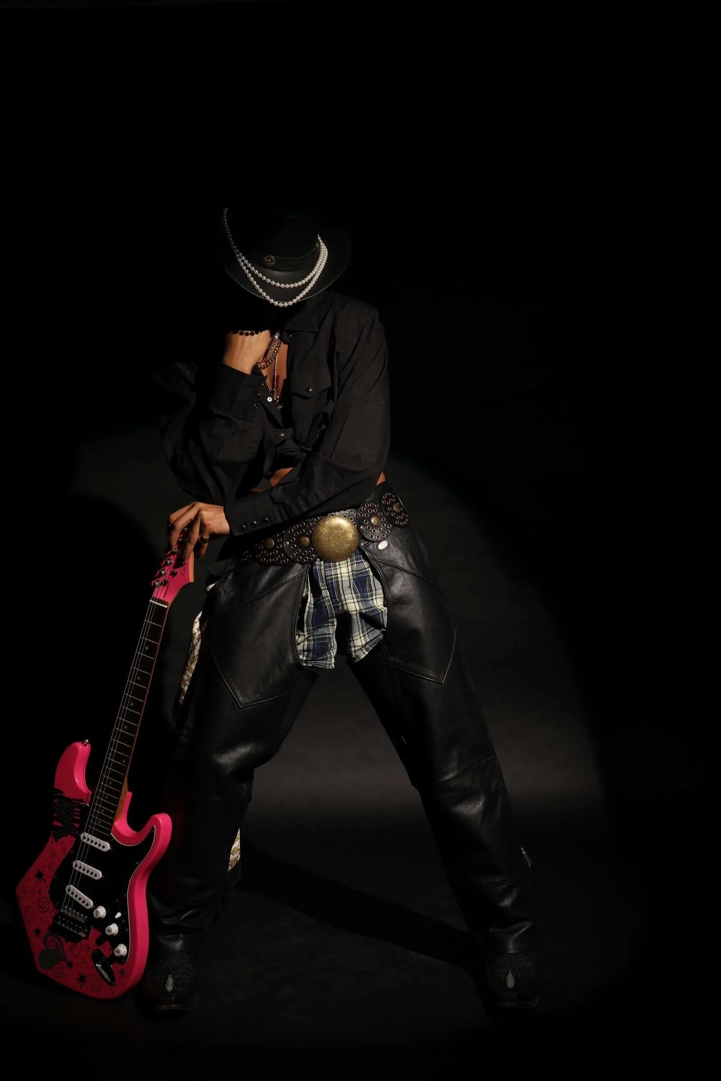Person dressed in black leather pants, black shirt, and accessories holding a pink electric guitar with black designs, against a dark background.