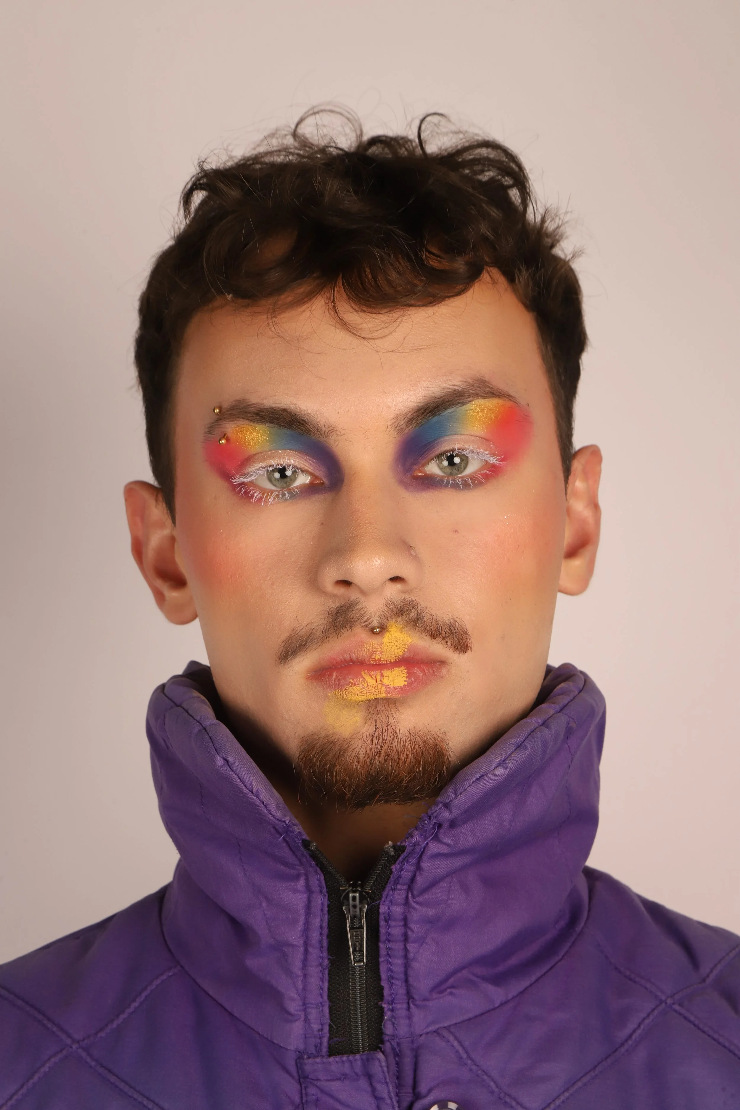 Person with rainbow colorful makeup, silver eye shadow, and a purple jacket, looking at the camera.