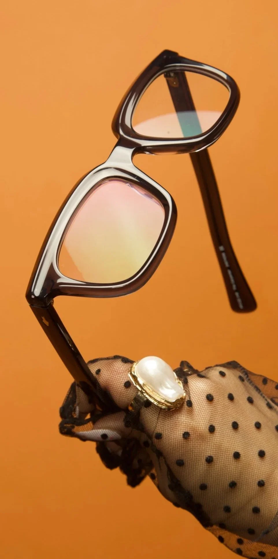 Fingertip with a black polka dot mesh glove holding sunglasses and a pearl ring against an orange background.