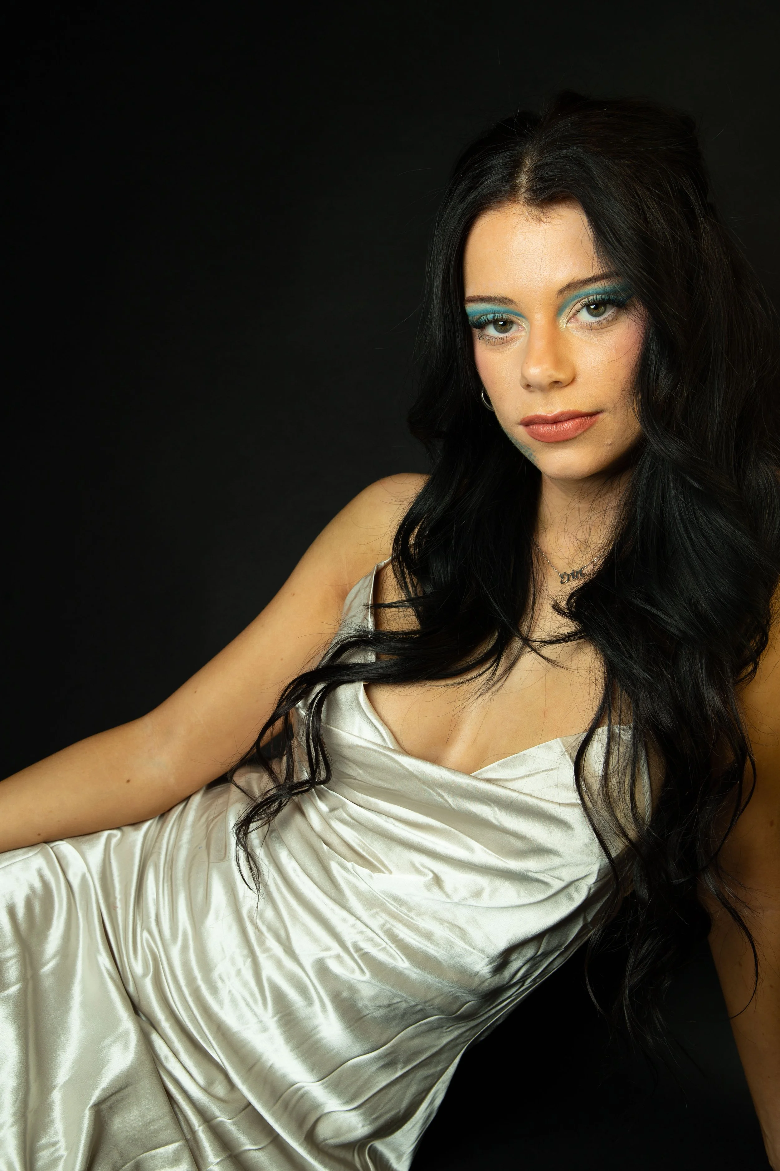 Young woman with black hair and blue eyeshadow wearing a shiny beige dress, posing against a black background.