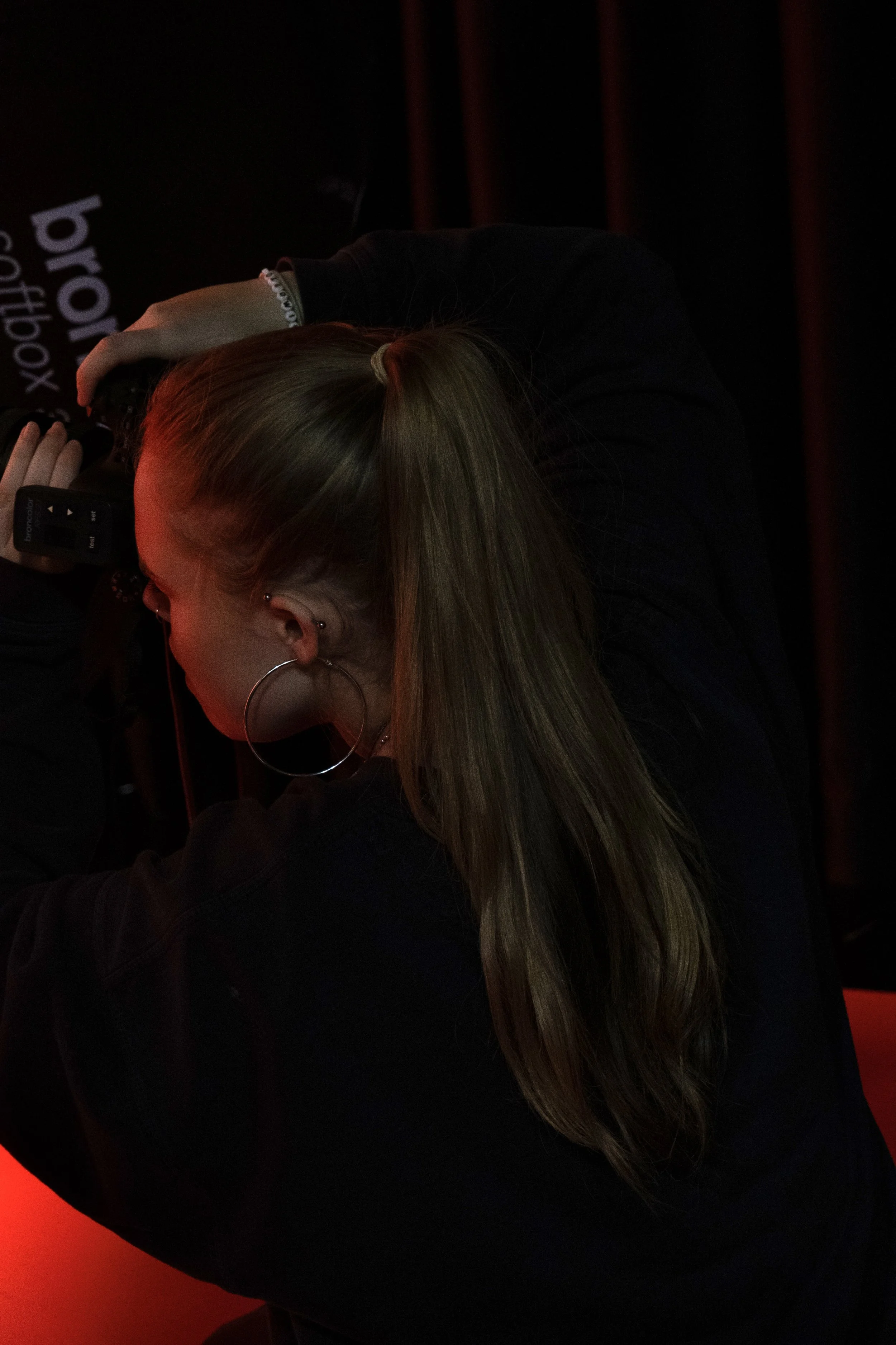 A woman with long hair tied back, wearing large hoop earrings and a black top, is in a dark room taking a photo with a camera.