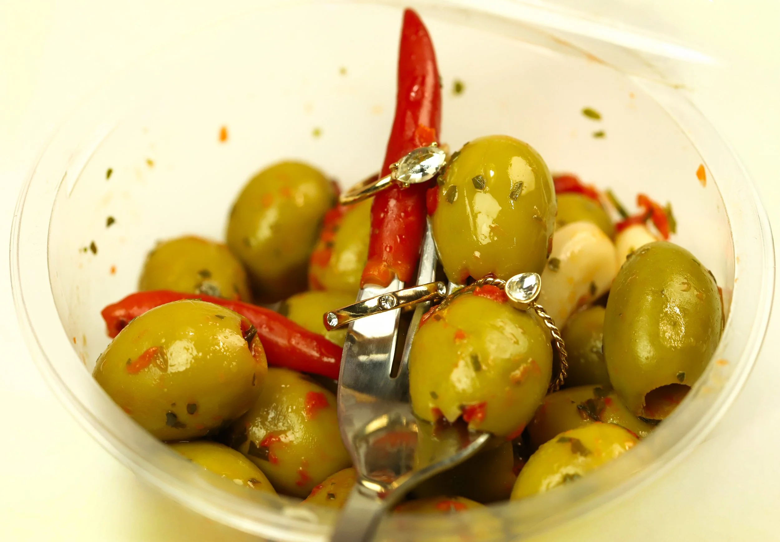 Green olives and red chili peppers in a transparent container, with a pair of silver jewelry pieces on top.
