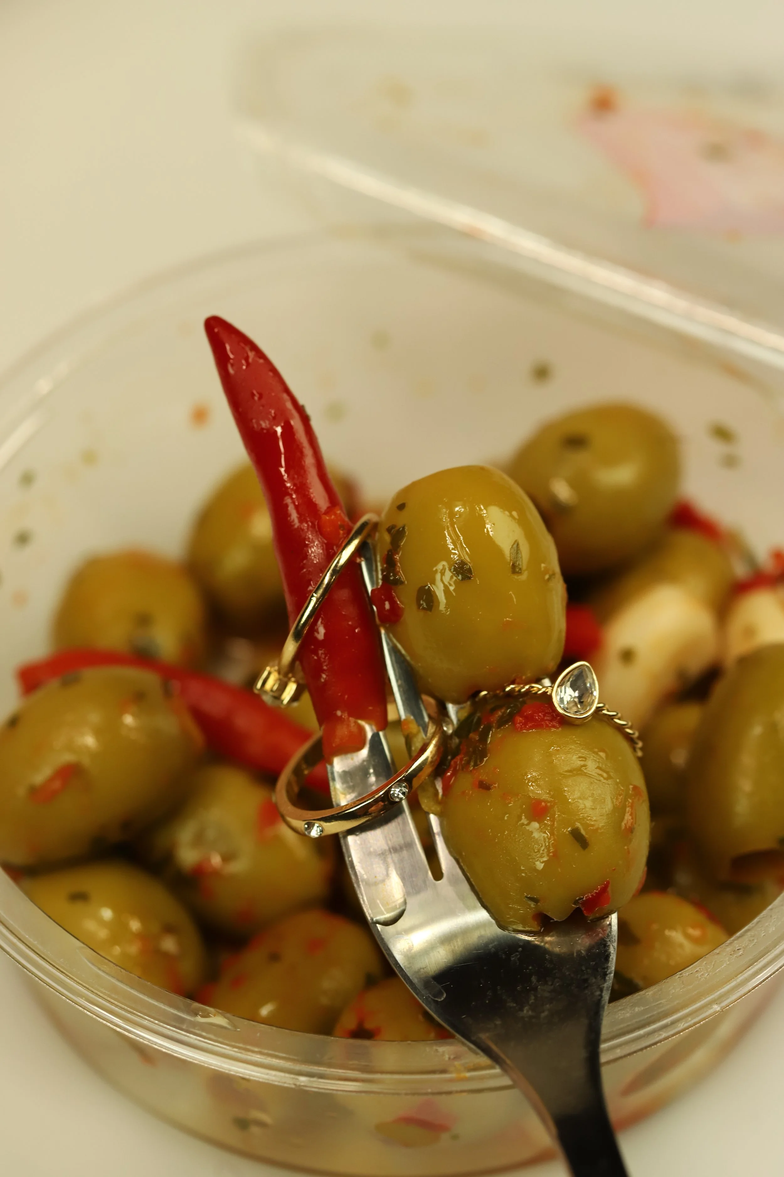 A fork holding pickled green olives and a red chili pepper, with more olives in a clear plastic container in the background.