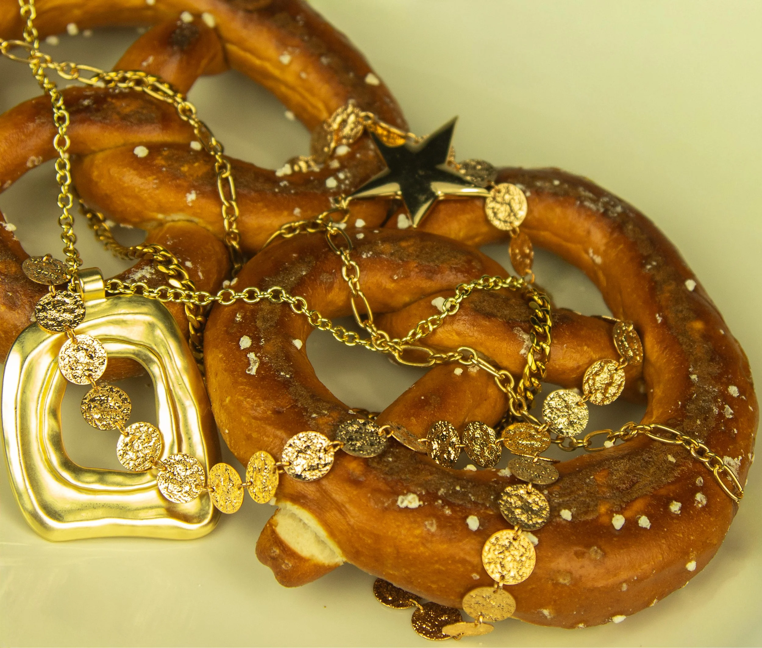 A salted pretzel twisted into a knot with gold jewelry, including necklaces, a star-shaped pendant, and a heart-shaped pendant, draped over it.