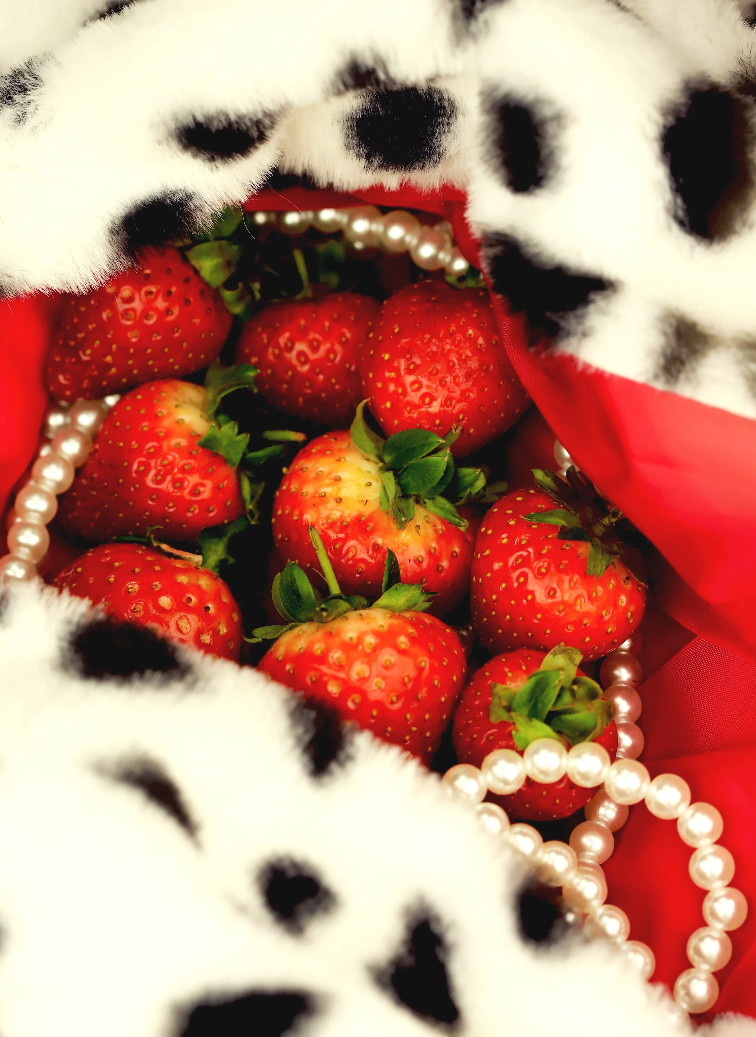 Fresh strawberries inside a red gift bag, decorated with a white faux fur with black spots and a string of white pearls.