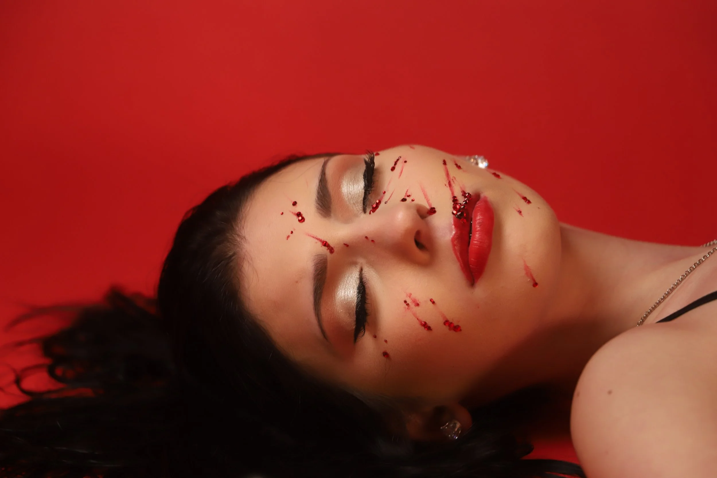 A woman with closed eyes lying down, with blood splatters on her face, on a red background.