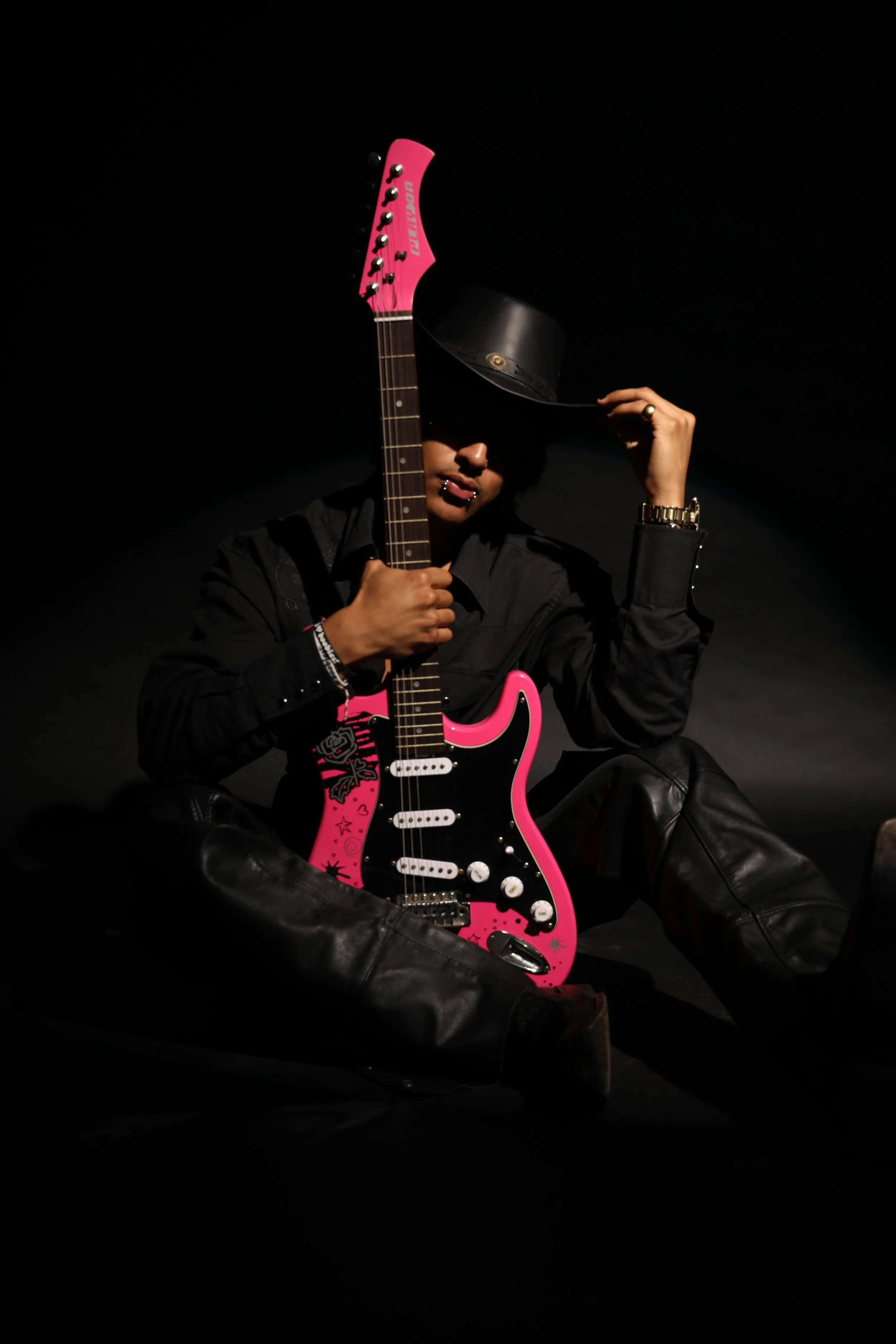 Person sitting with legs crossed, holding a pink electric guitar, wearing a black hat and black clothing, in a dark setting.