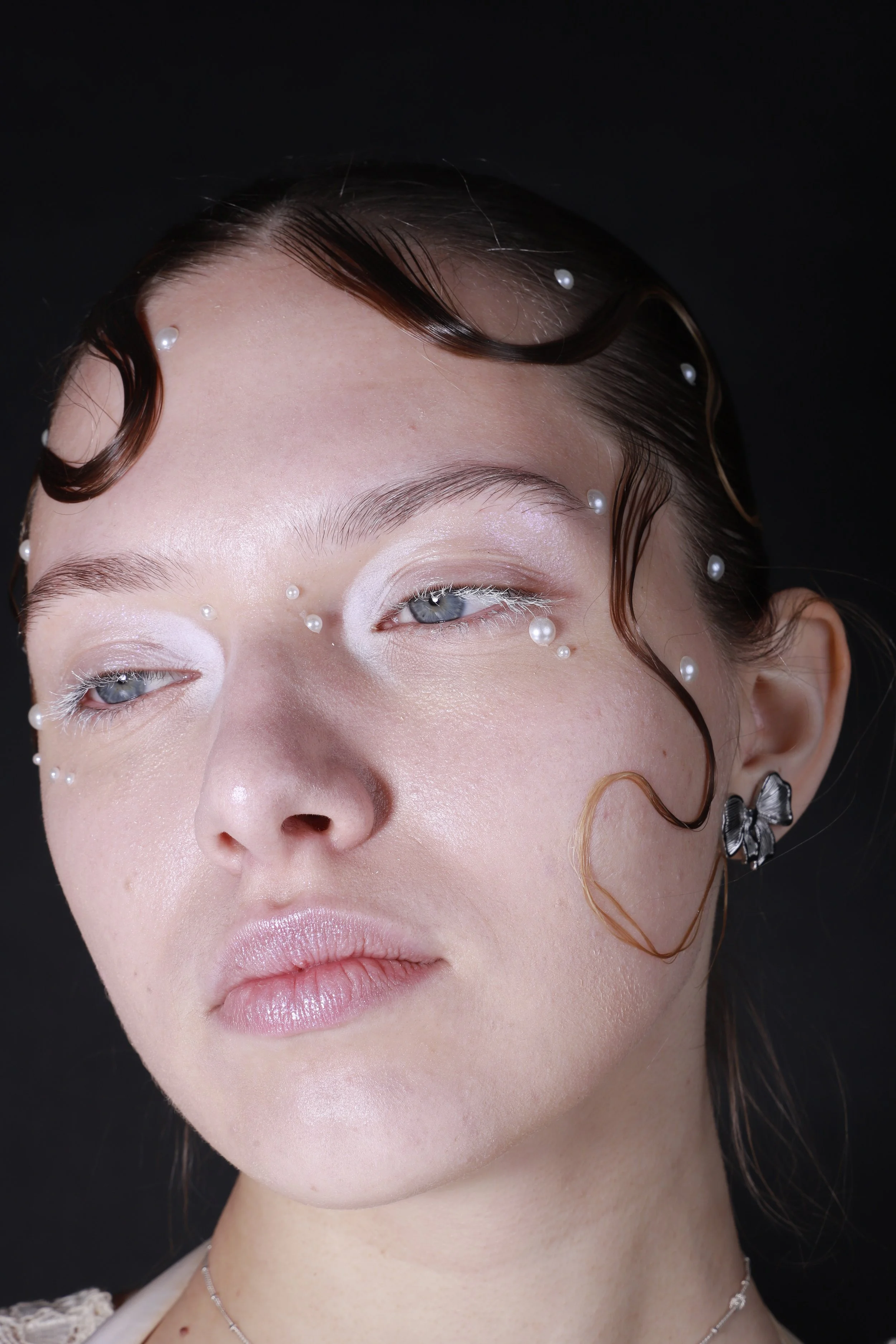 Close-up of a woman's face with pale skin, light makeup, pale pink lips, and blue eyes. Her dark brown hair is styled with wavy strands, some decorated with pearls, and she wears butterfly-shaped earrings.
