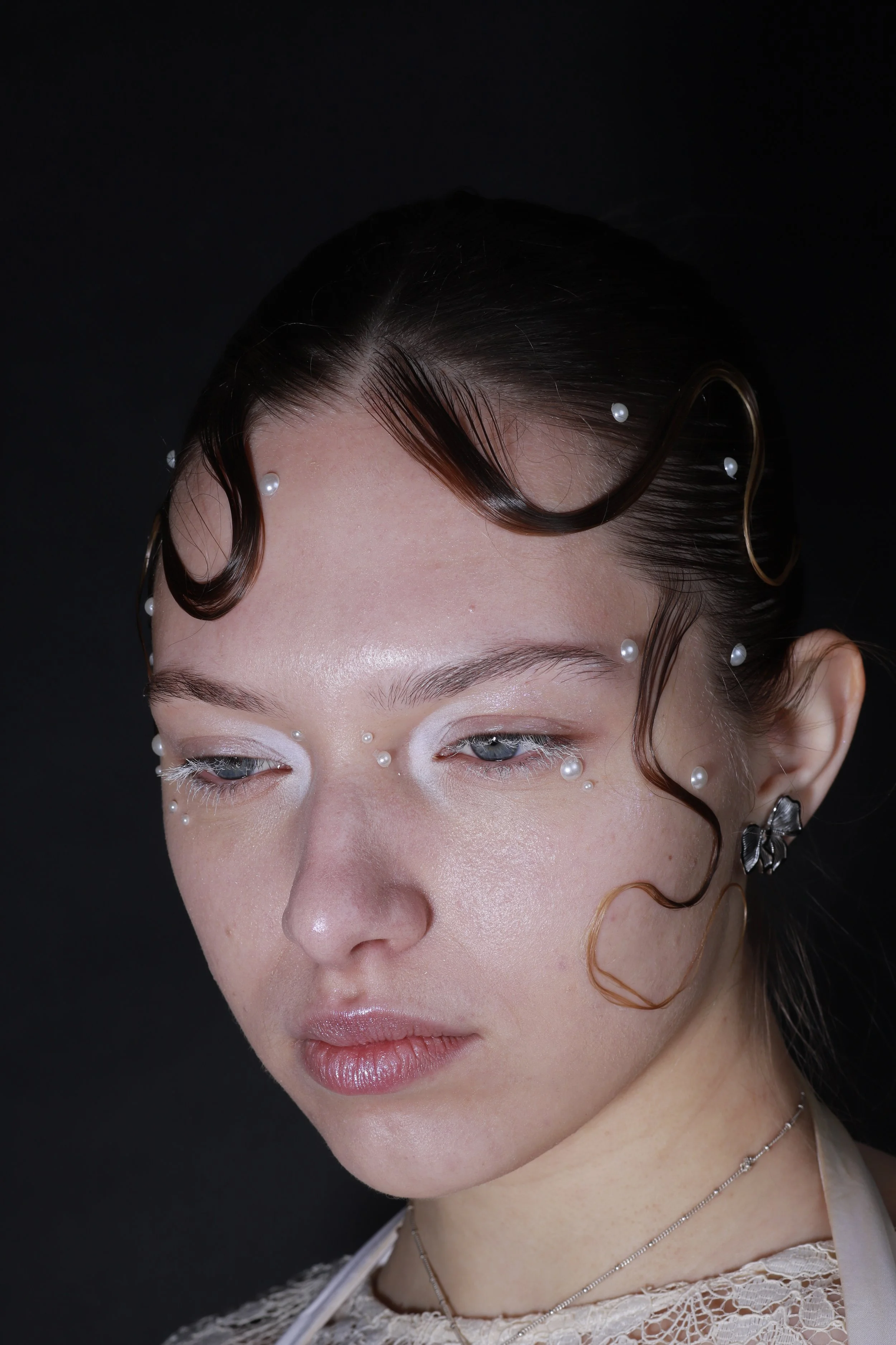 A close-up of a woman with styled hair and pearl and rhinestone accessories on her face and head, against a black background.