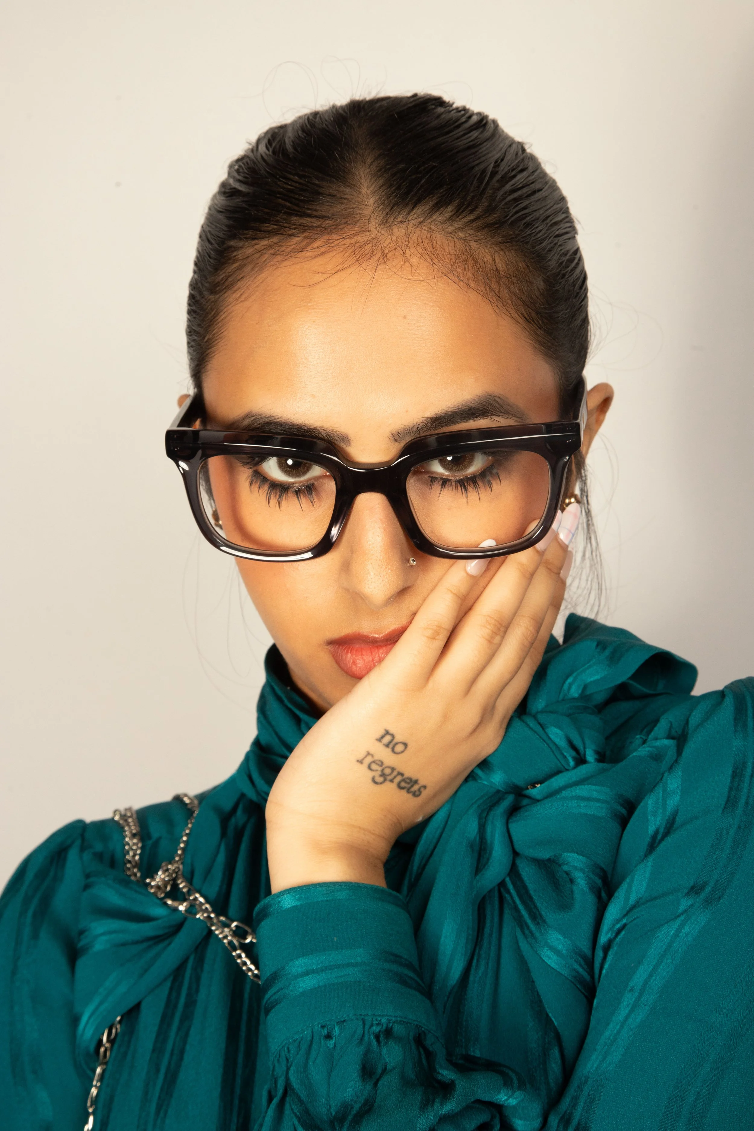 A woman with dark hair in a bun, wearing big black glasses, a teal jacket with a chain, and a tattoo on her hand that says 'no regrets', posing with her hand near her face.