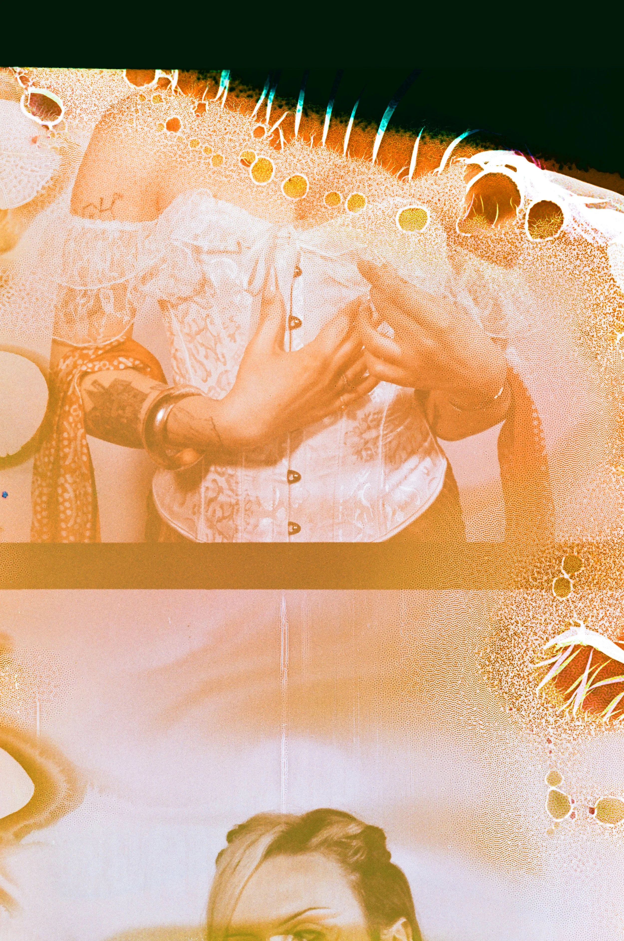 Close-up of a person folding a white shirt with visible tattoos on their arms, jewelry, and rings, with a person with blond hair partially visible at the bottom.