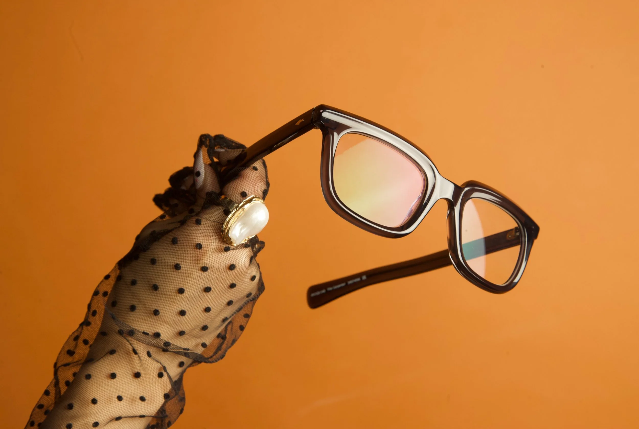 A hand wearing a sheer, black polka-dotted glove holds a pair of black-rimmed glasses with tinted lenses against a solid orange background.