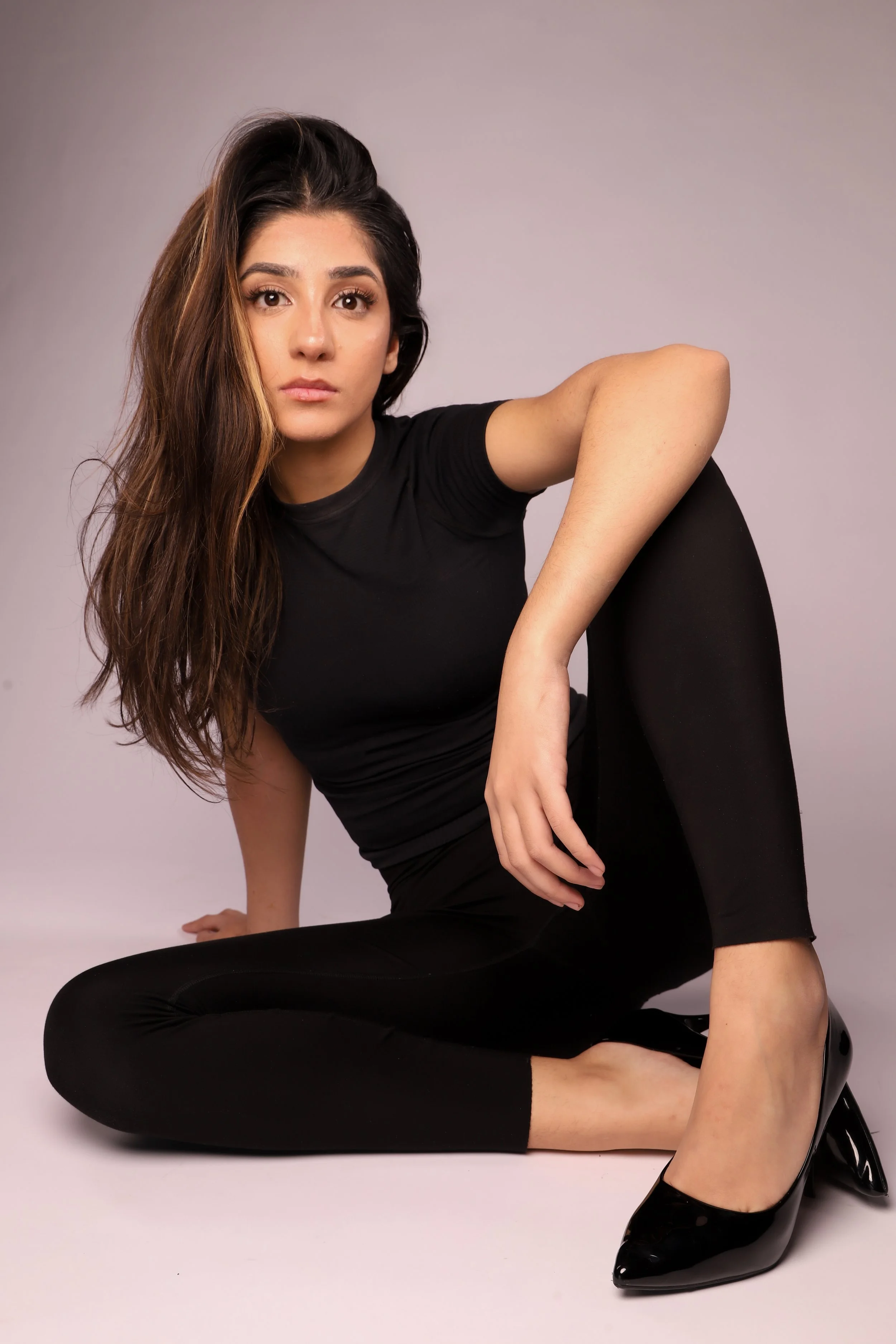 A woman with long, wavy brown hair and light skin wearing a black t-shirt, black leggings, and black high heels, sitting on the floor against a plain light background.