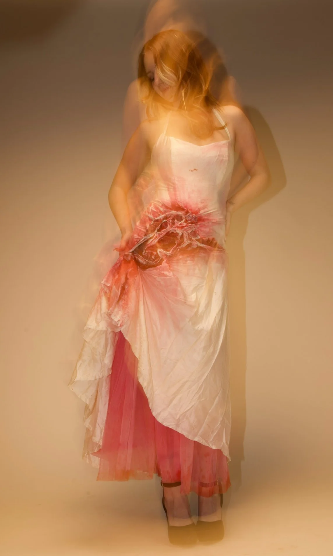 A woman with long red hair wearing a long, flowing dress with pink, white, and red colors, standing against a plain background. The image appears to have a motion blur or double exposure effect.
