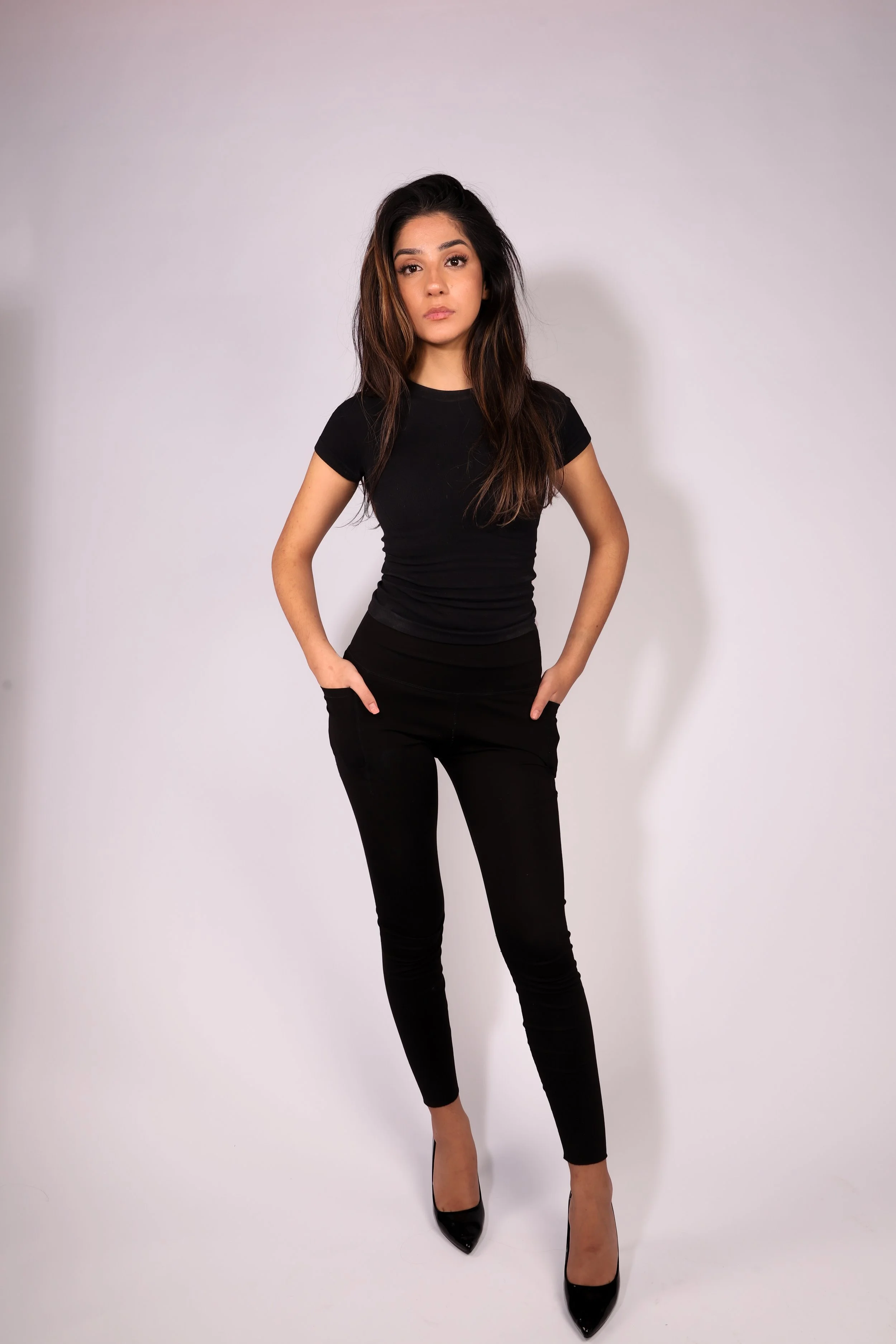 A young woman with long dark hair, wearing a black t-shirt, black pants, and black high heels, standing against a plain light gray background.