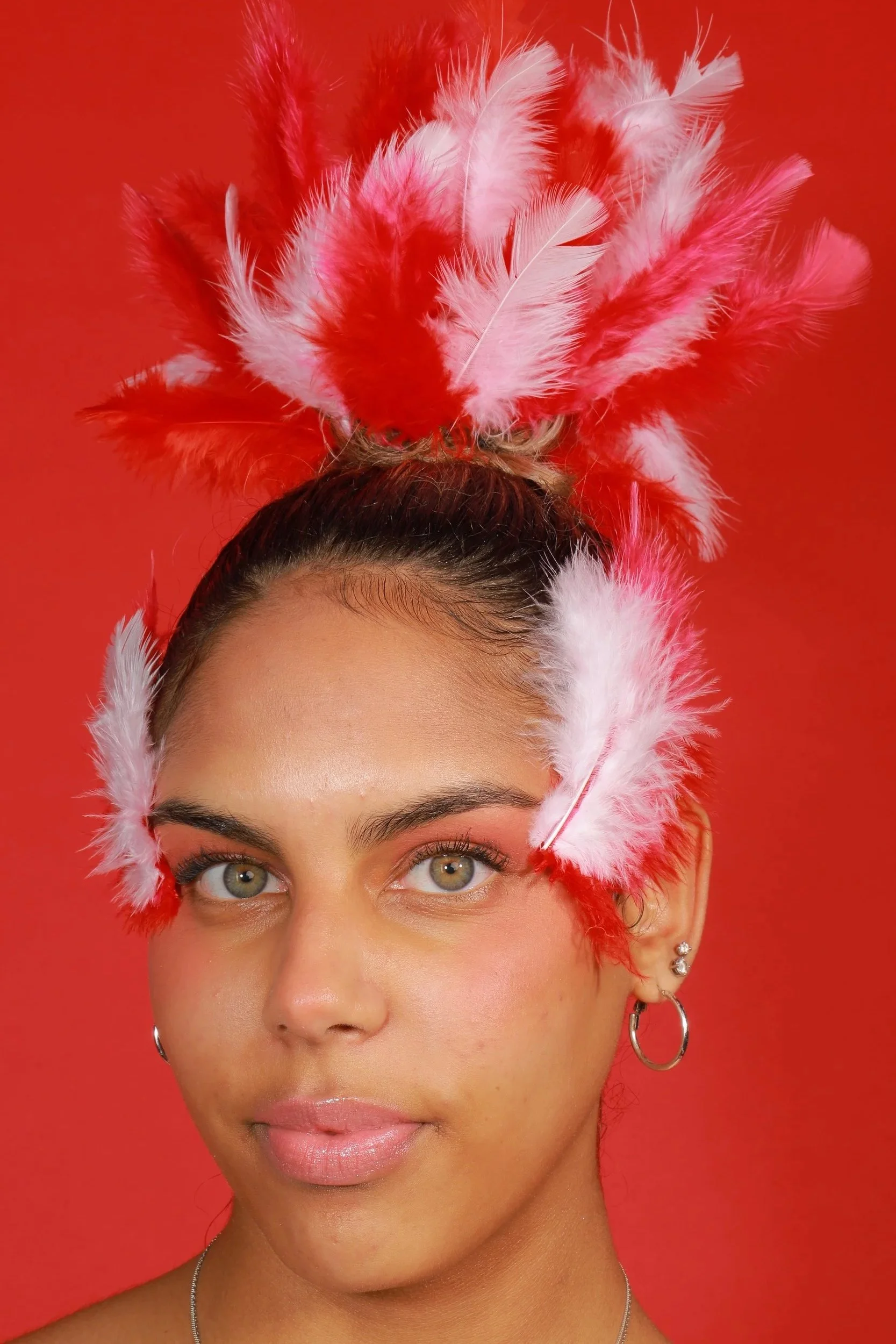 A young woman with light-colored eyes wearing pink and white feather accessories on her face and hair against a red background.