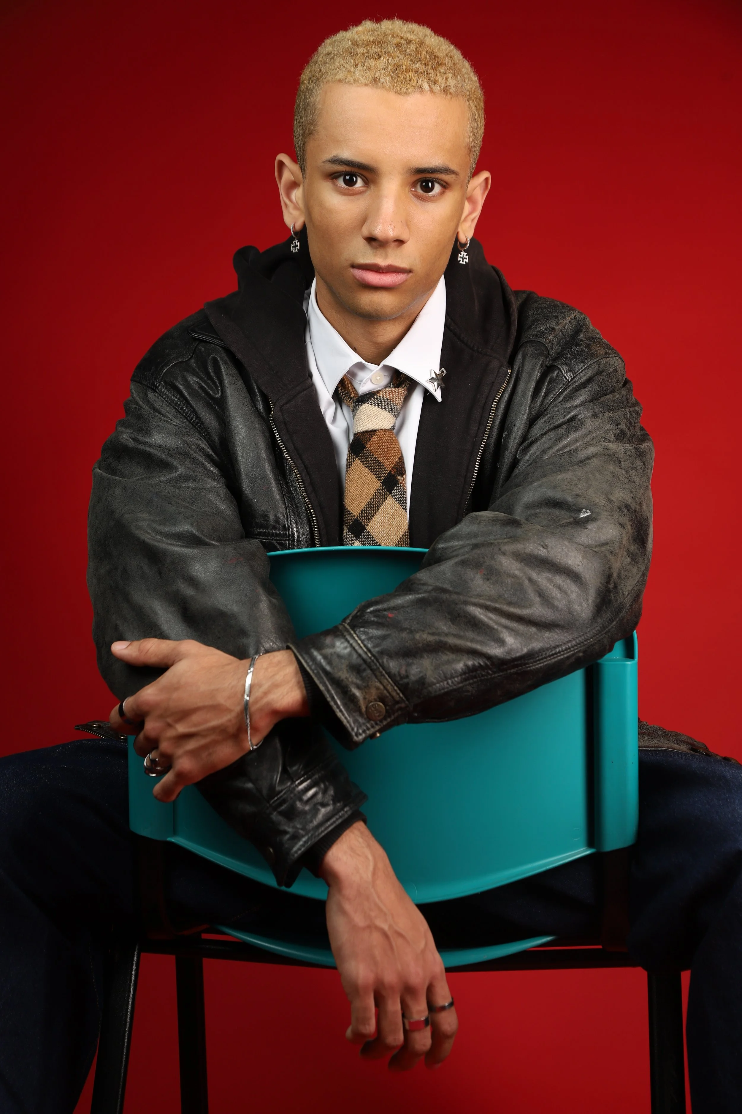 Young person with short blond hair, wearing a white shirt, a plaid tie, a black jacket, earrings, and rings, sitting on a chair with a red background.