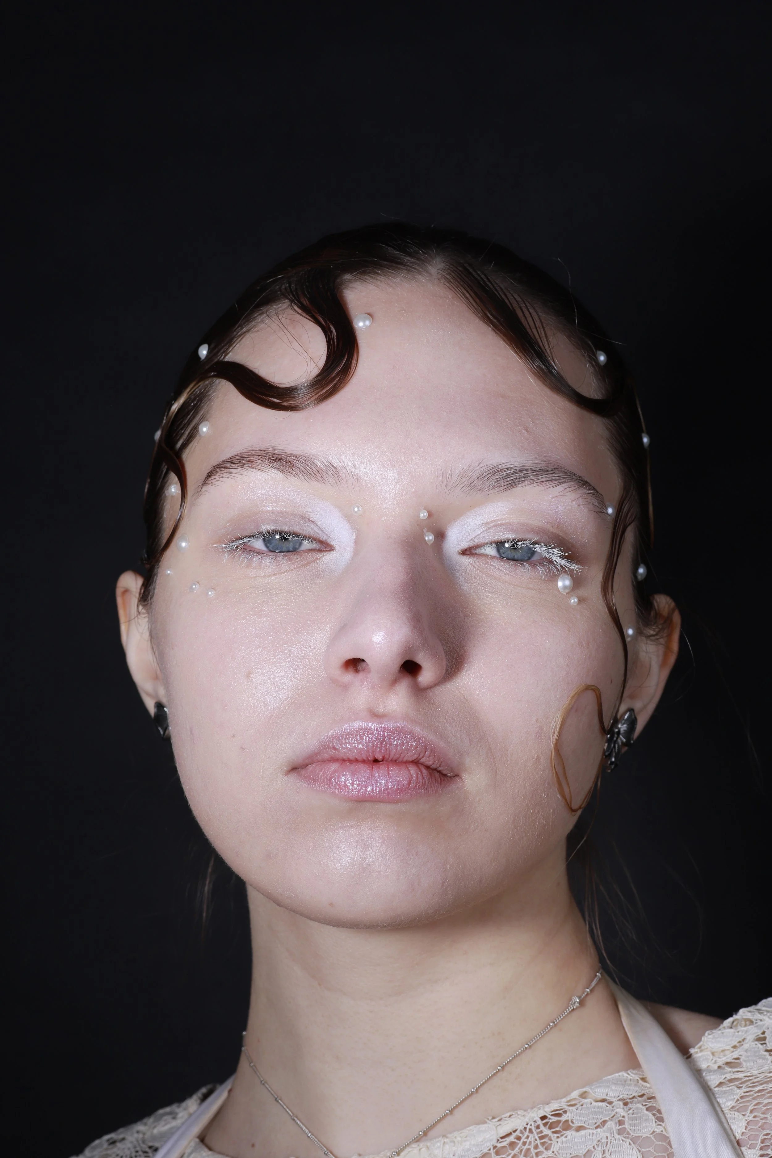 Close-up of a woman with wet hair decorated with pearls, wearing light makeup, against a black background.