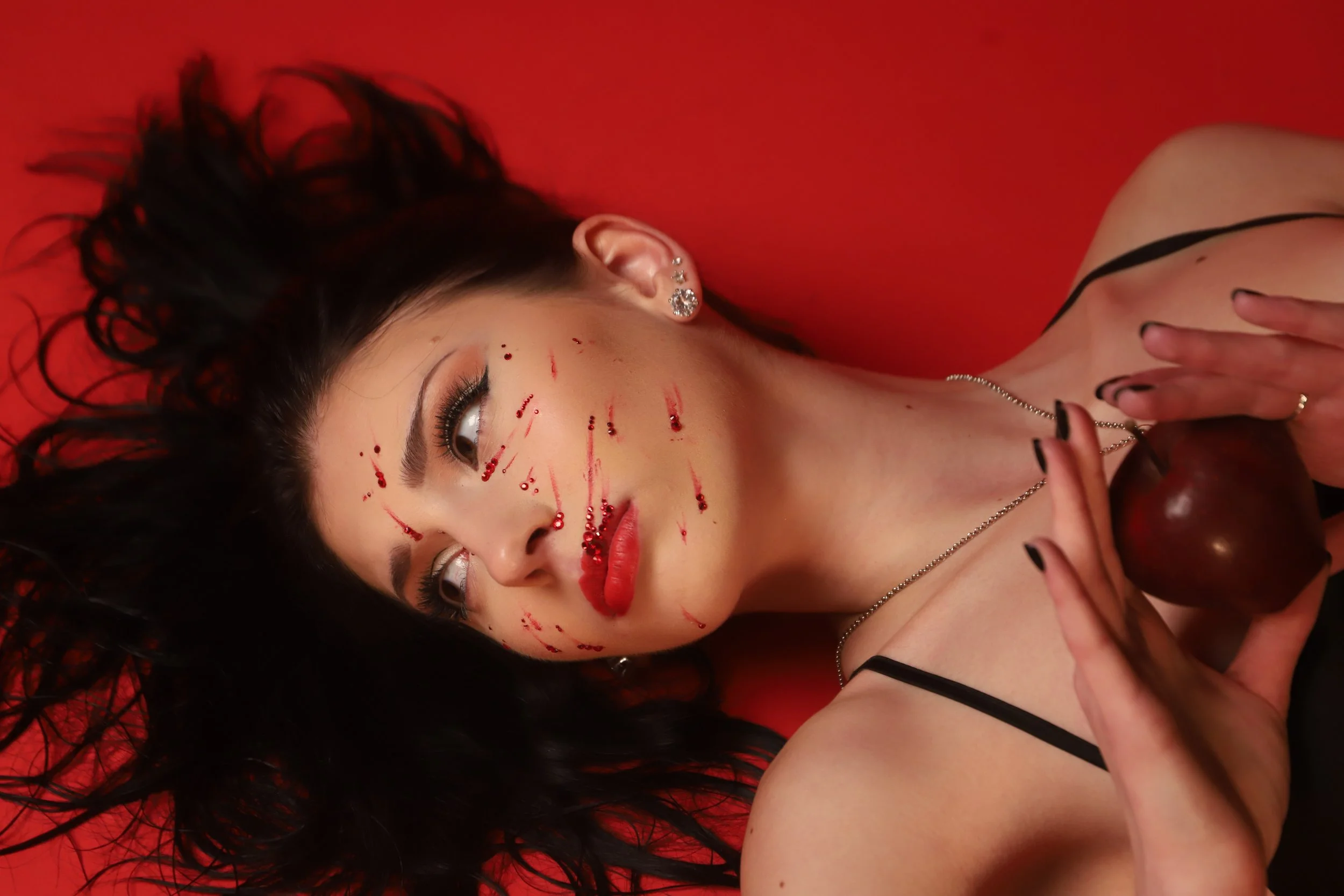 A woman with dark, wavy hair lying on a red background, holding a cherry close to her chest, with blood-like streaks on her face and makeup resembling scars.