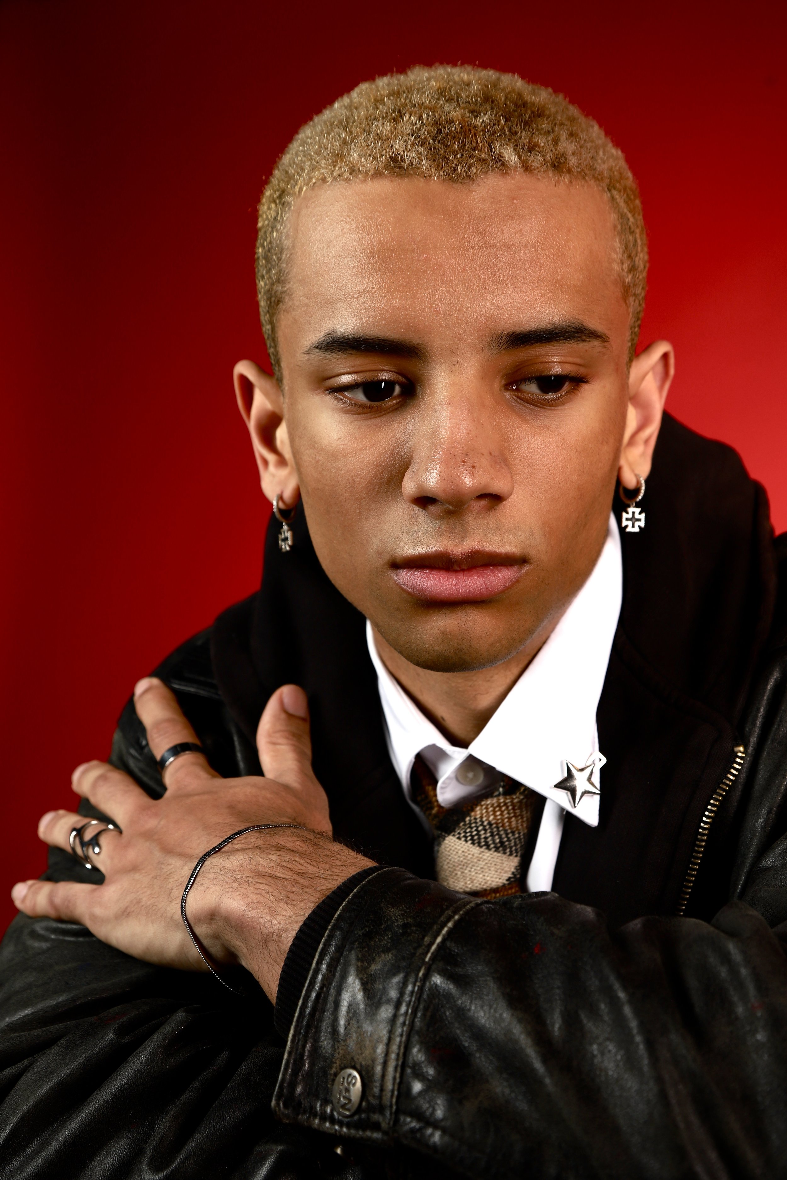 Portrait of a young man with short blonde hair, wearing a black leather jacket, white shirt, and a patterned tie, with earrings and rings, posing against a red background.