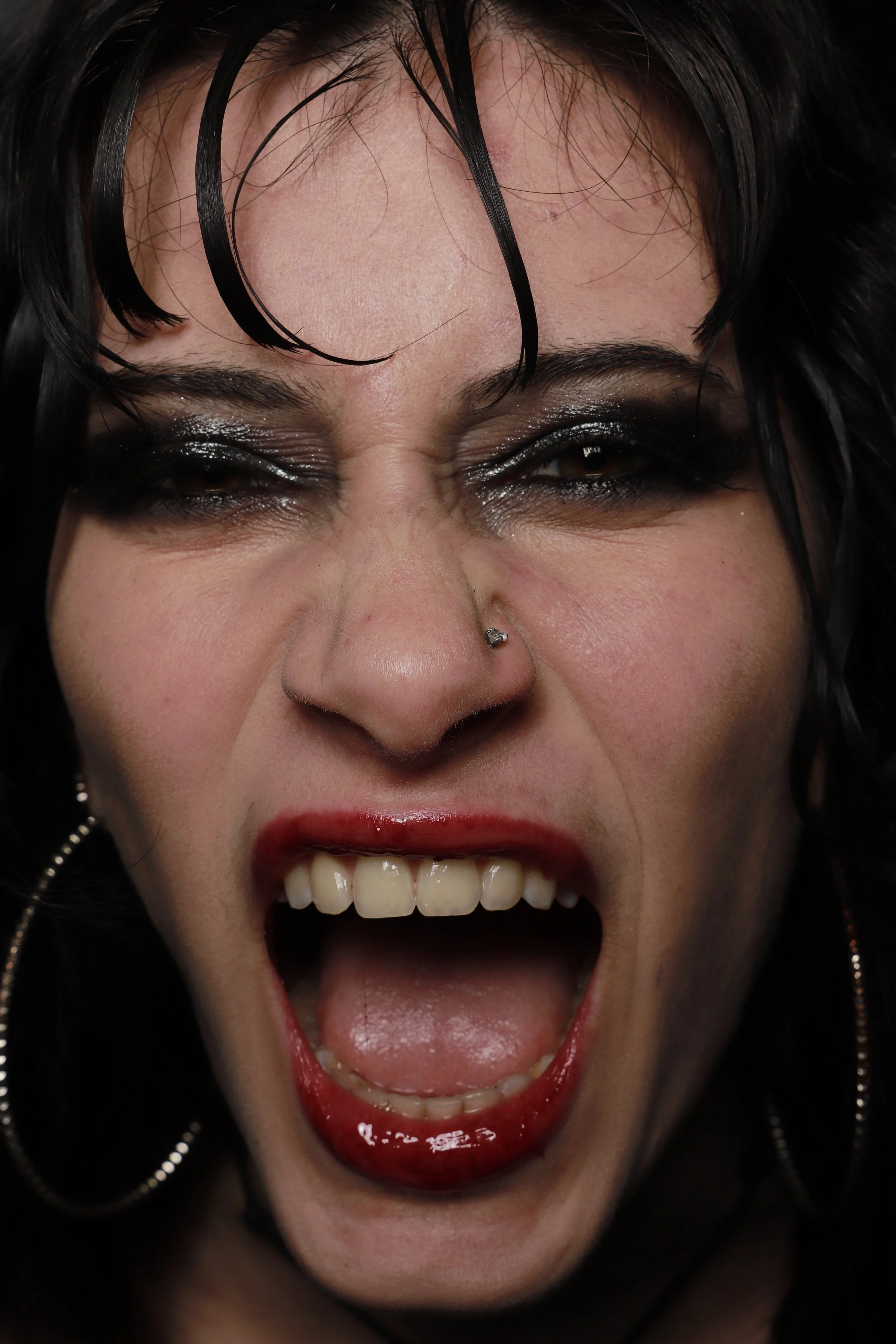 Close-up of a woman with dark hair, black makeup, and red lipstick, screaming with her mouth wide open.