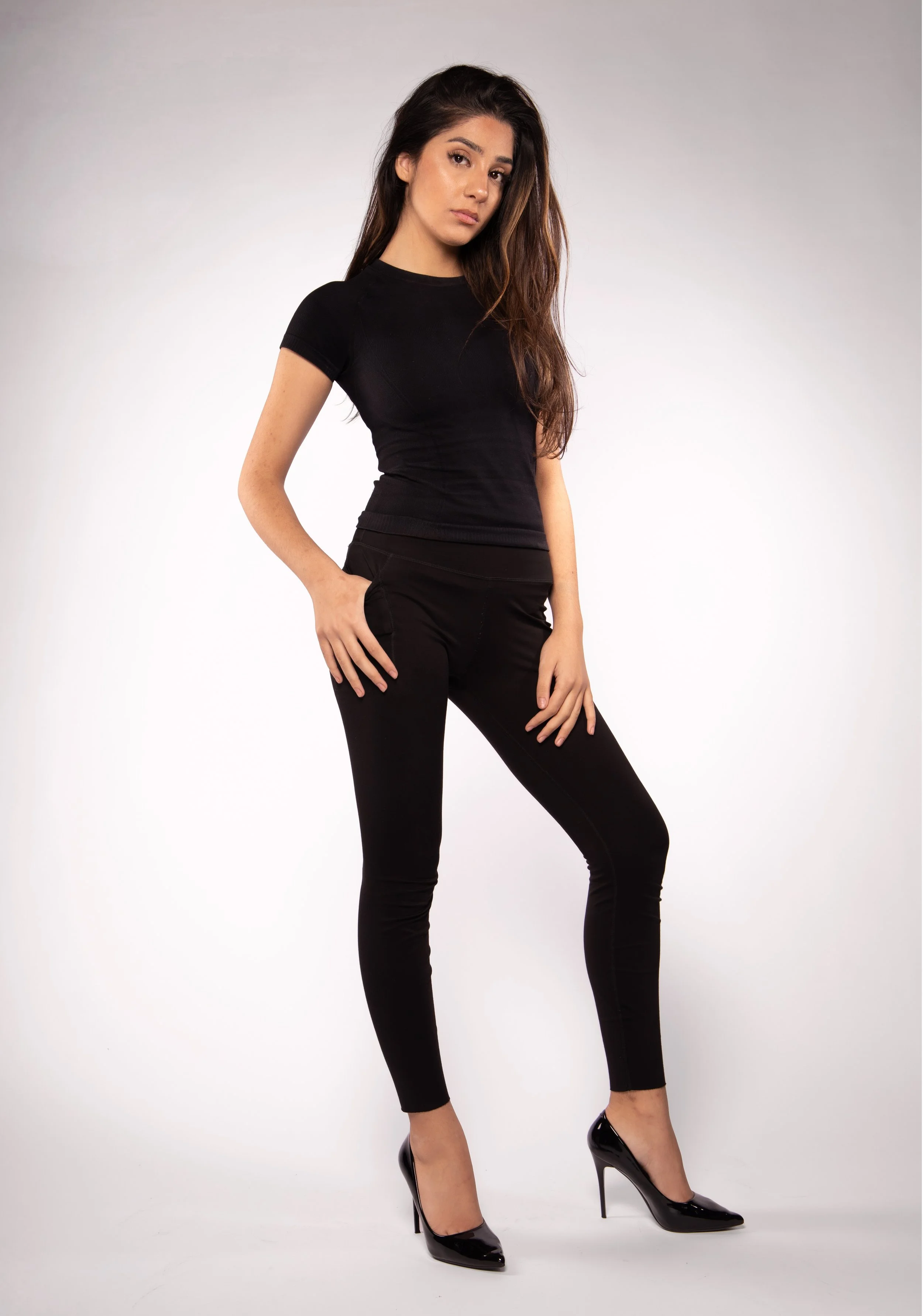 A young woman with long, wavy brown hair in black attire, black high heels, standing against a plain background.