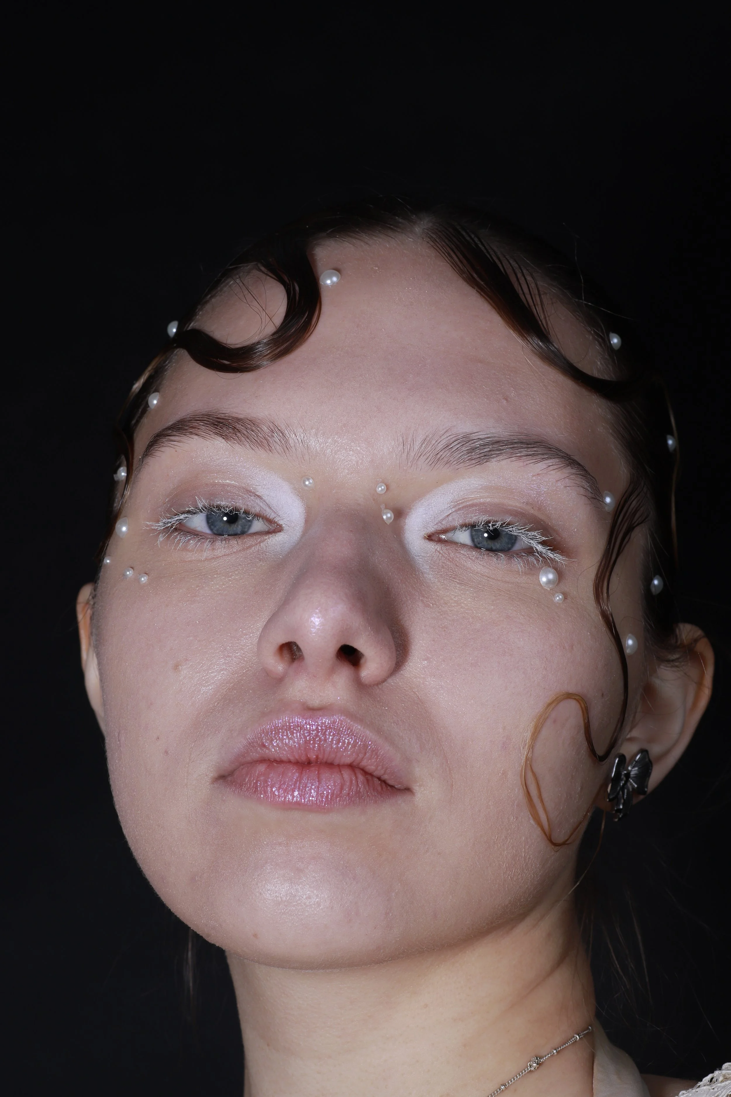 Close-up of a woman with blue eyes and light makeup, adorned with small pearl-like beads on her face and hair, against a black background.