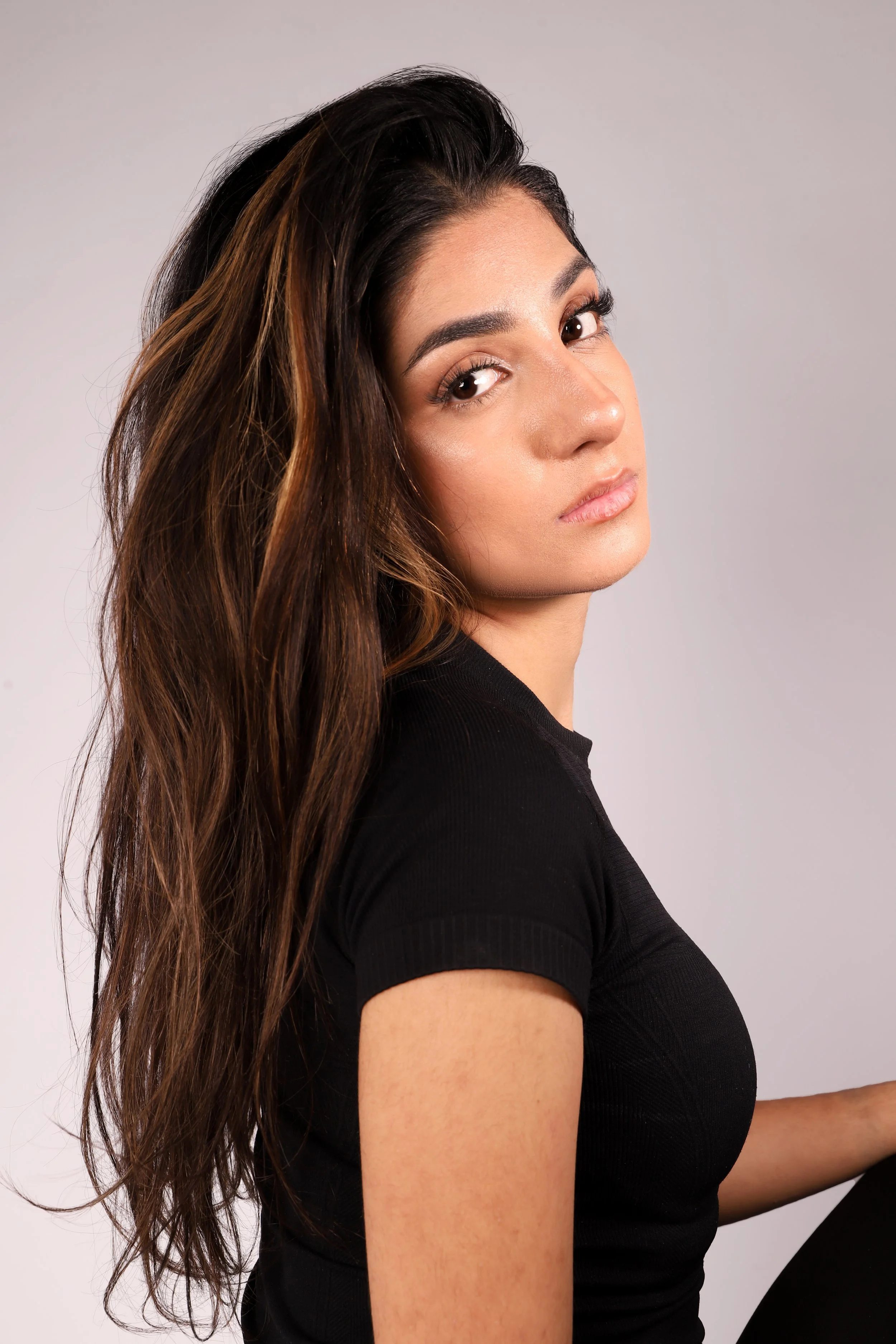 A young woman with long, wavy brown hair and light skin looking at the camera, wearing a black top, against a light gray background.