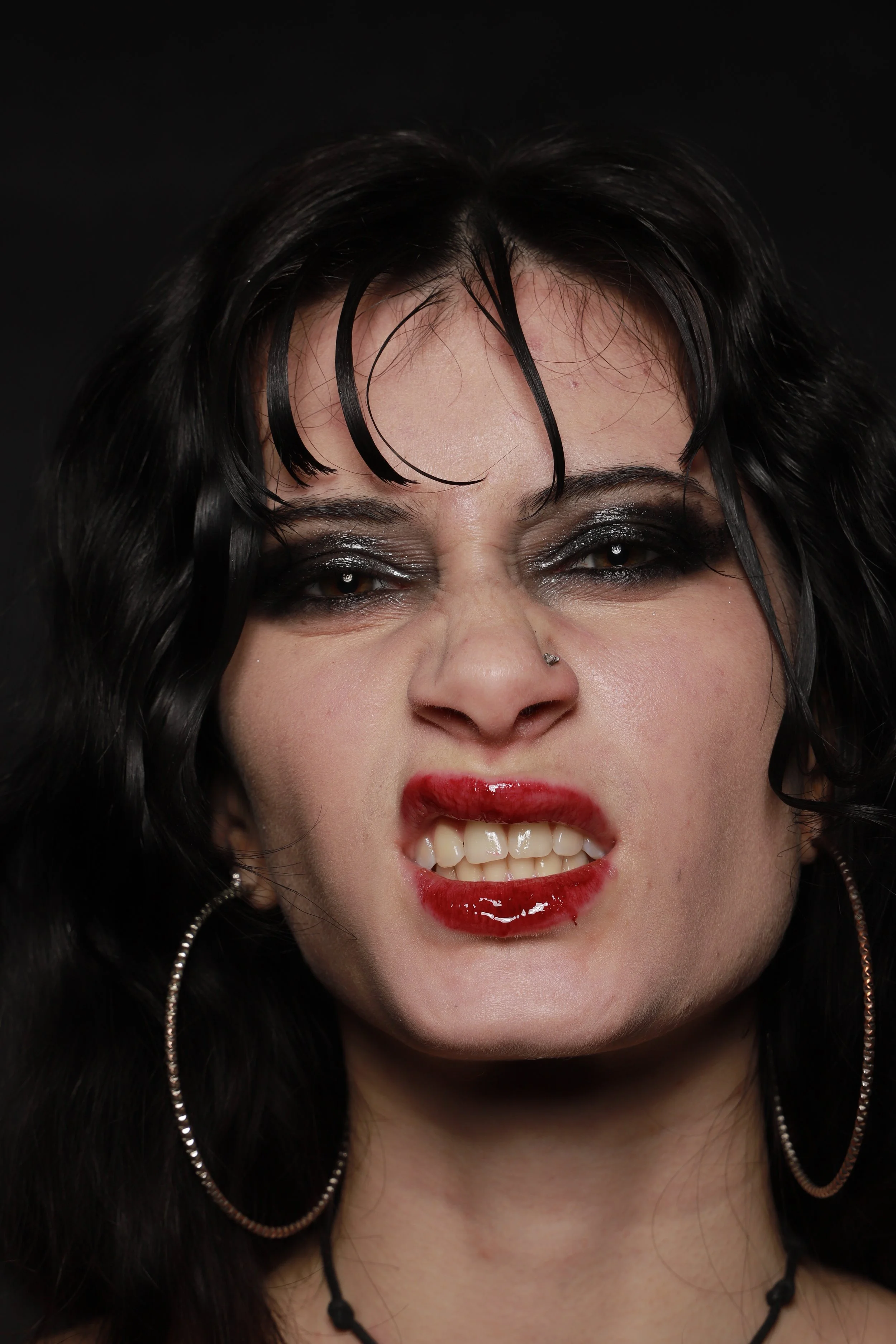 Close-up of a woman with black, wavy hair, dark eye makeup, red lipstick, lip gloss, hoop earrings, a small nose piercing, and an angry or fierce expression, showing clenched teeth and furrowed brows against a black background.
