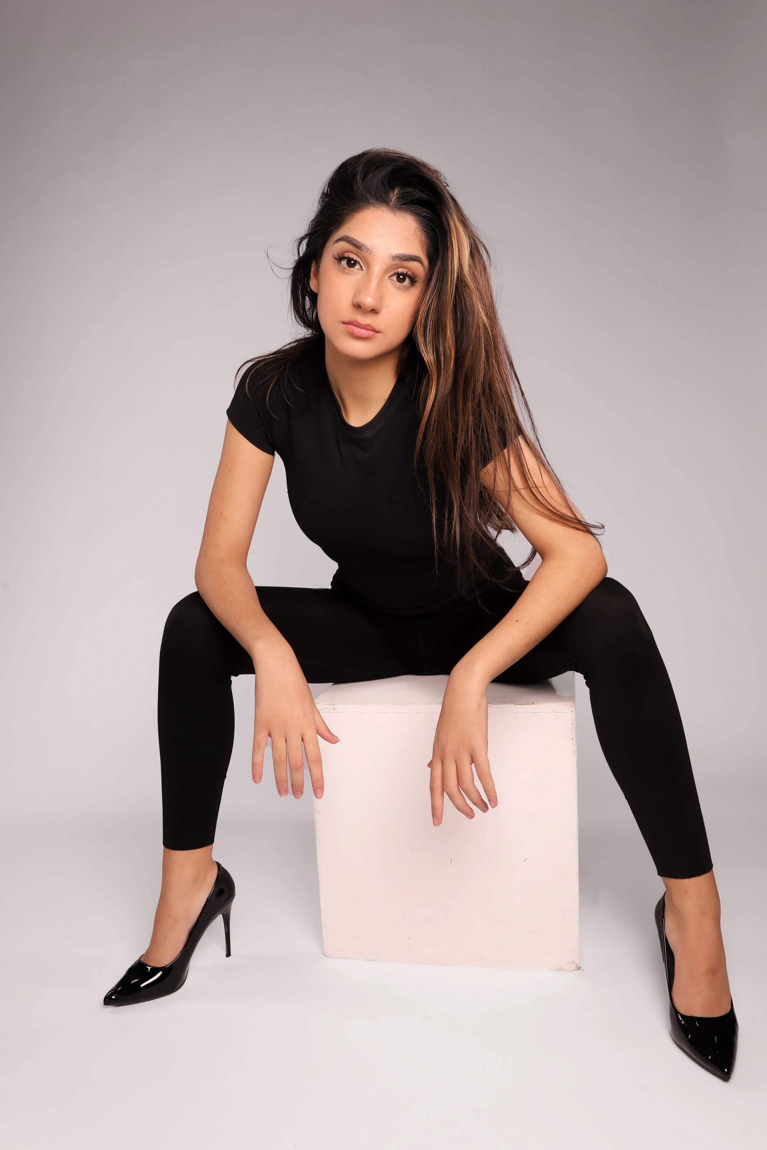 Young woman with long brown hair wearing a black t-shirt, black pants, and black high heels, sitting on a white cube against a gray background.