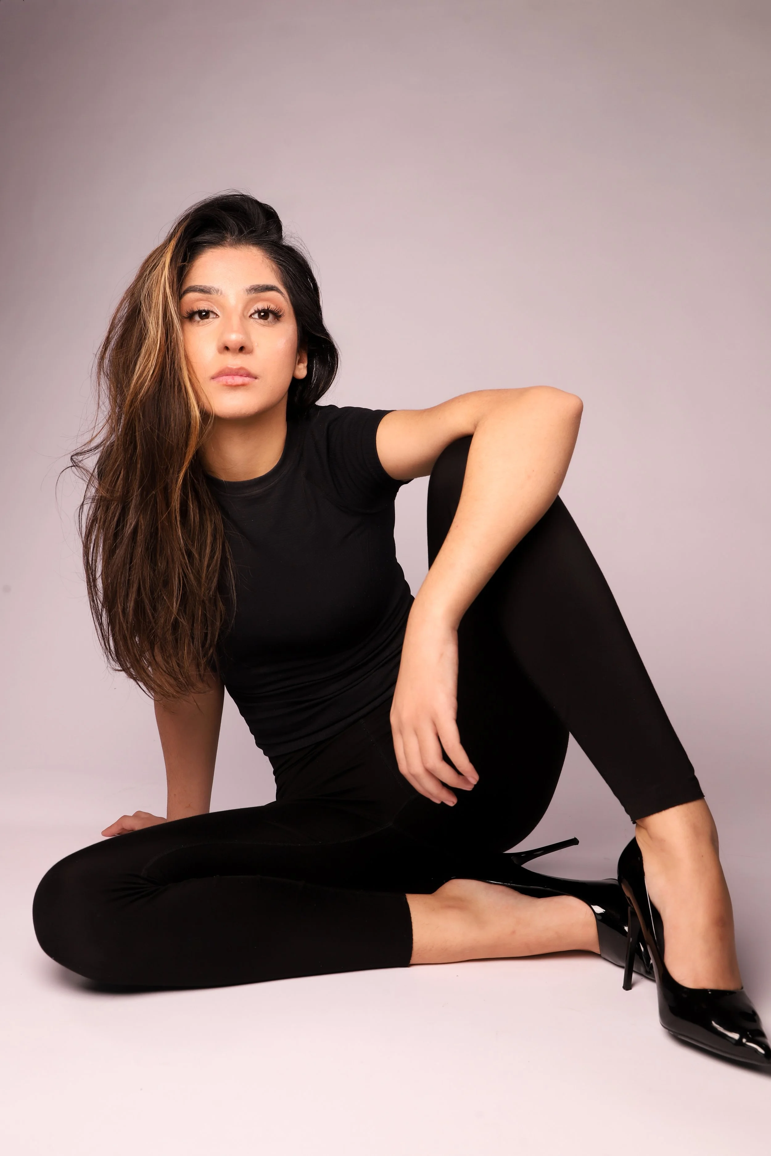 Young woman in black outfit and high heels sitting on the floor with a neutral background.