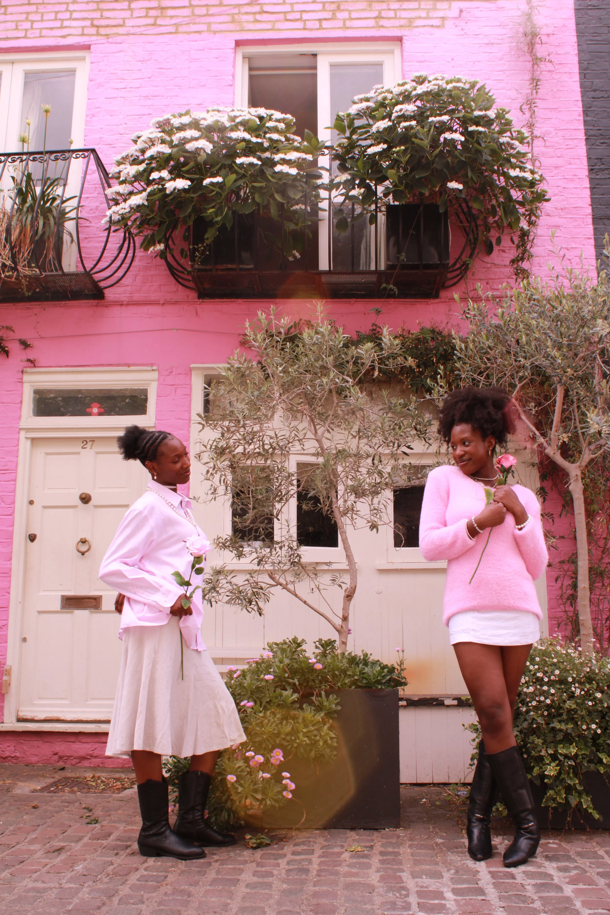 Two women standing on a cobblestone street in front of a pink building with potted plants and flowers. Both women are holding pink roses and wearing stylish outfits with boots, one in a light pink sweater and skirt, the other in a white blouse and skirt, smiling at each other.
