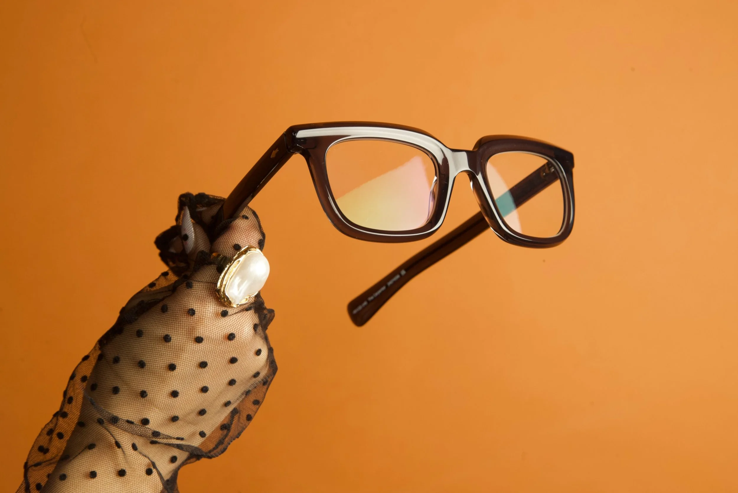 A hand wearing a polka dot mesh glove holding a pair of black-framed sunglasses with reflective lenses against an orange background.