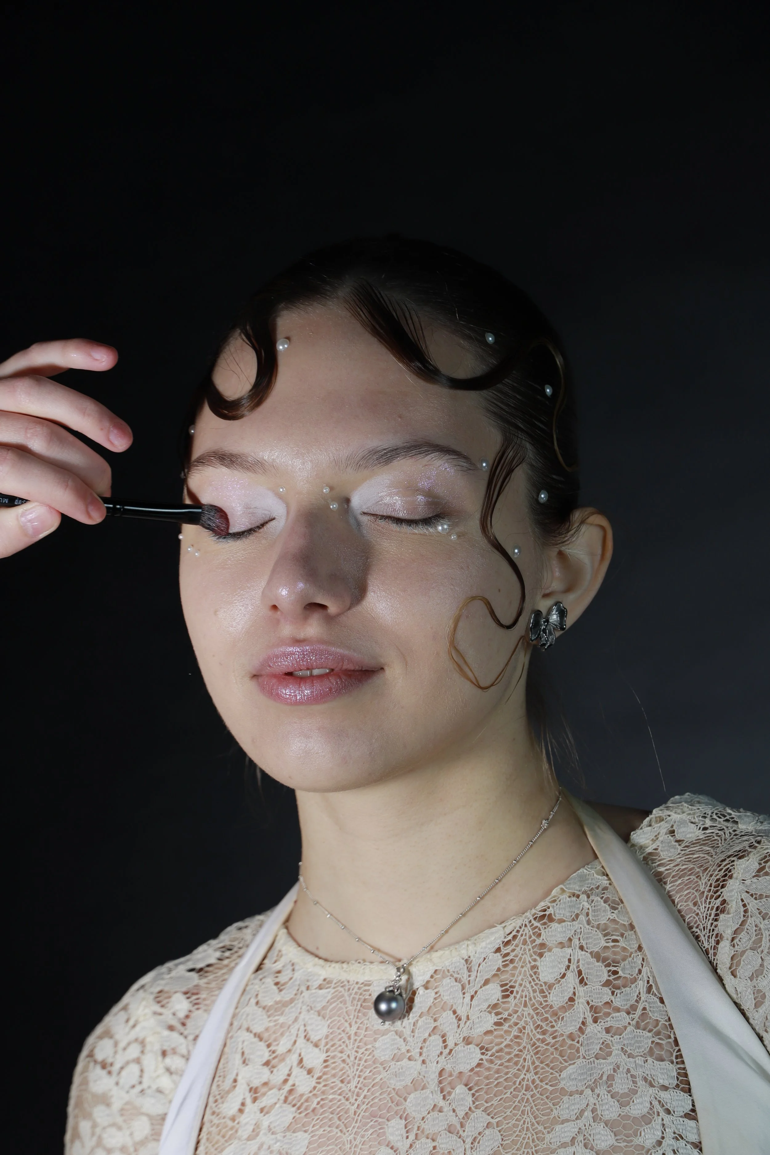 A woman with pearl embellishments in her hair and face is having makeup applied to her eyelids, against a dark background.