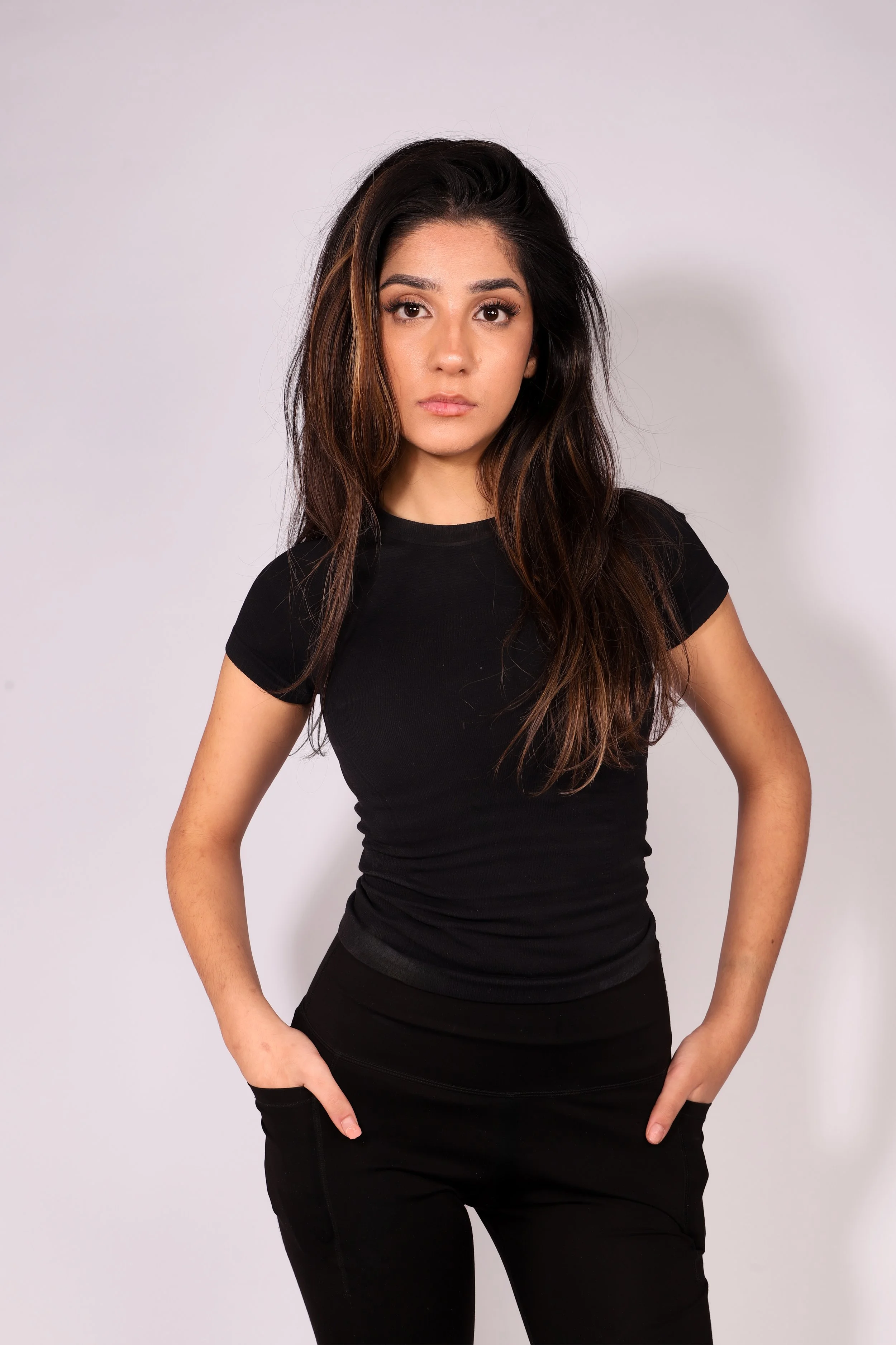 A young woman with long, wavy dark hair wearing a black t-shirt and black pants, standing against a plain light-colored background.