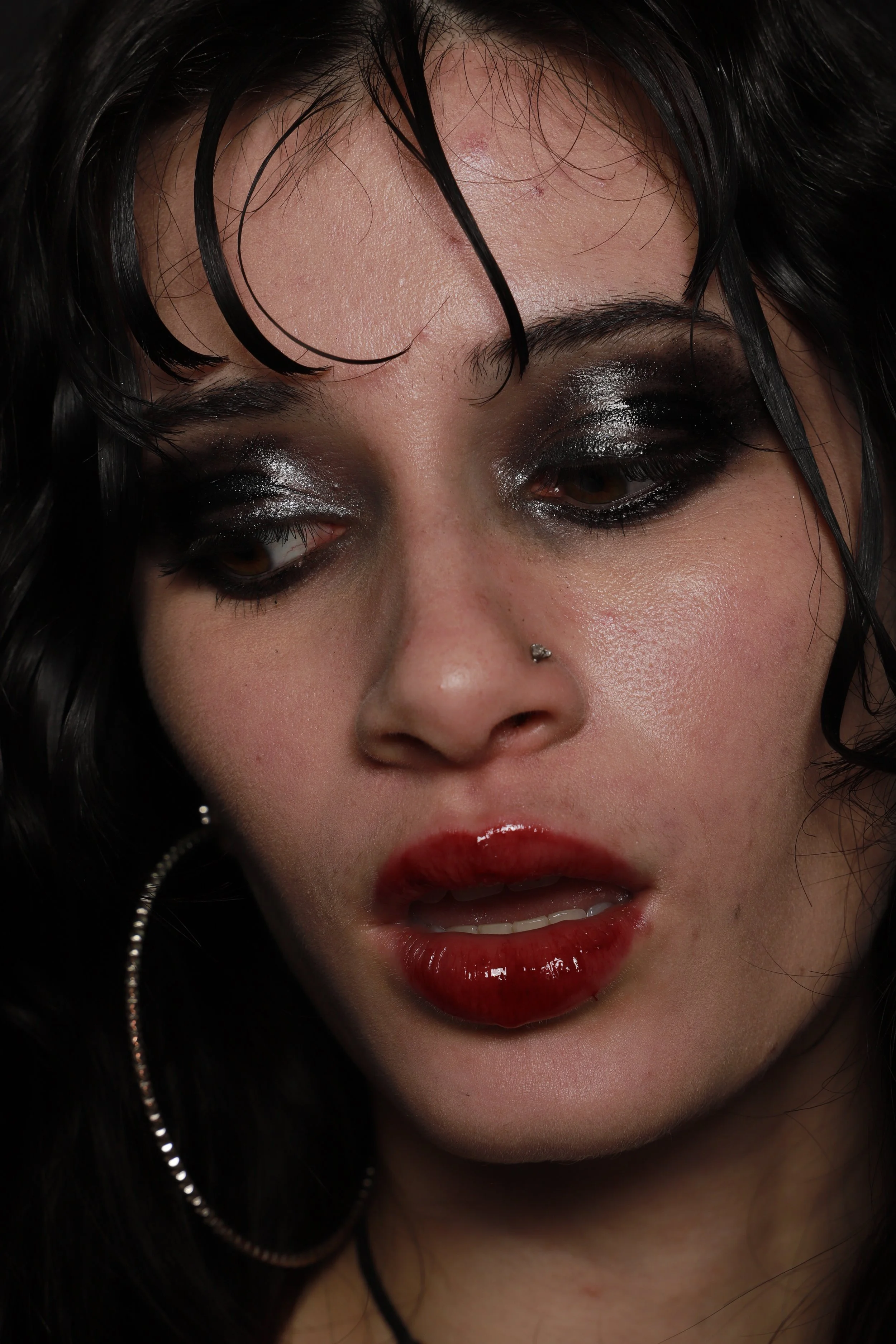 Close-up of a woman with dark, wavy hair, heavy black eye makeup, red lipstick, a nose piercing, and a large hoop earring.