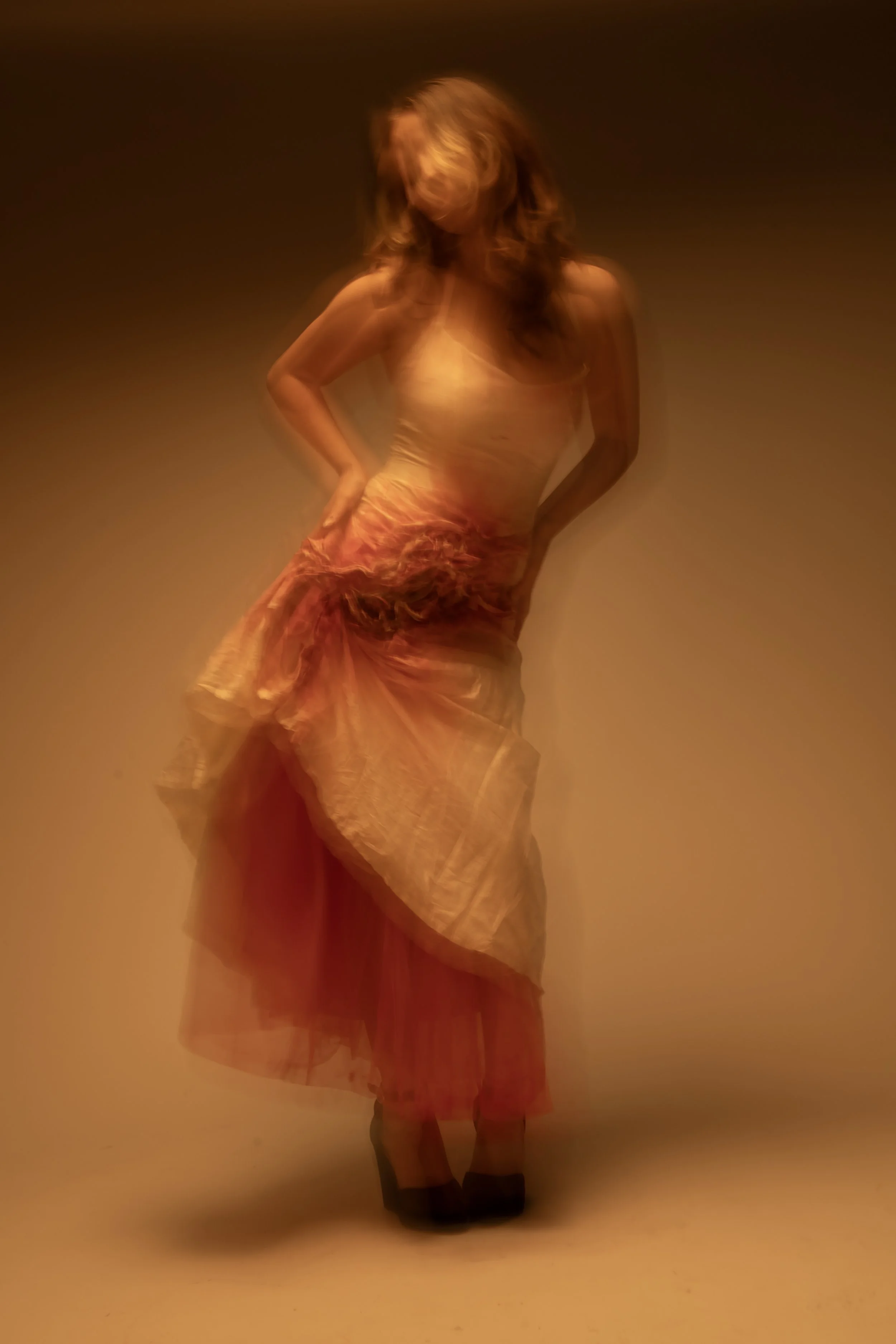 A woman with red hair wearing a flowing, semi-transparent orange and pink dress, standing against a plain background, with a blurred, artistic effect.