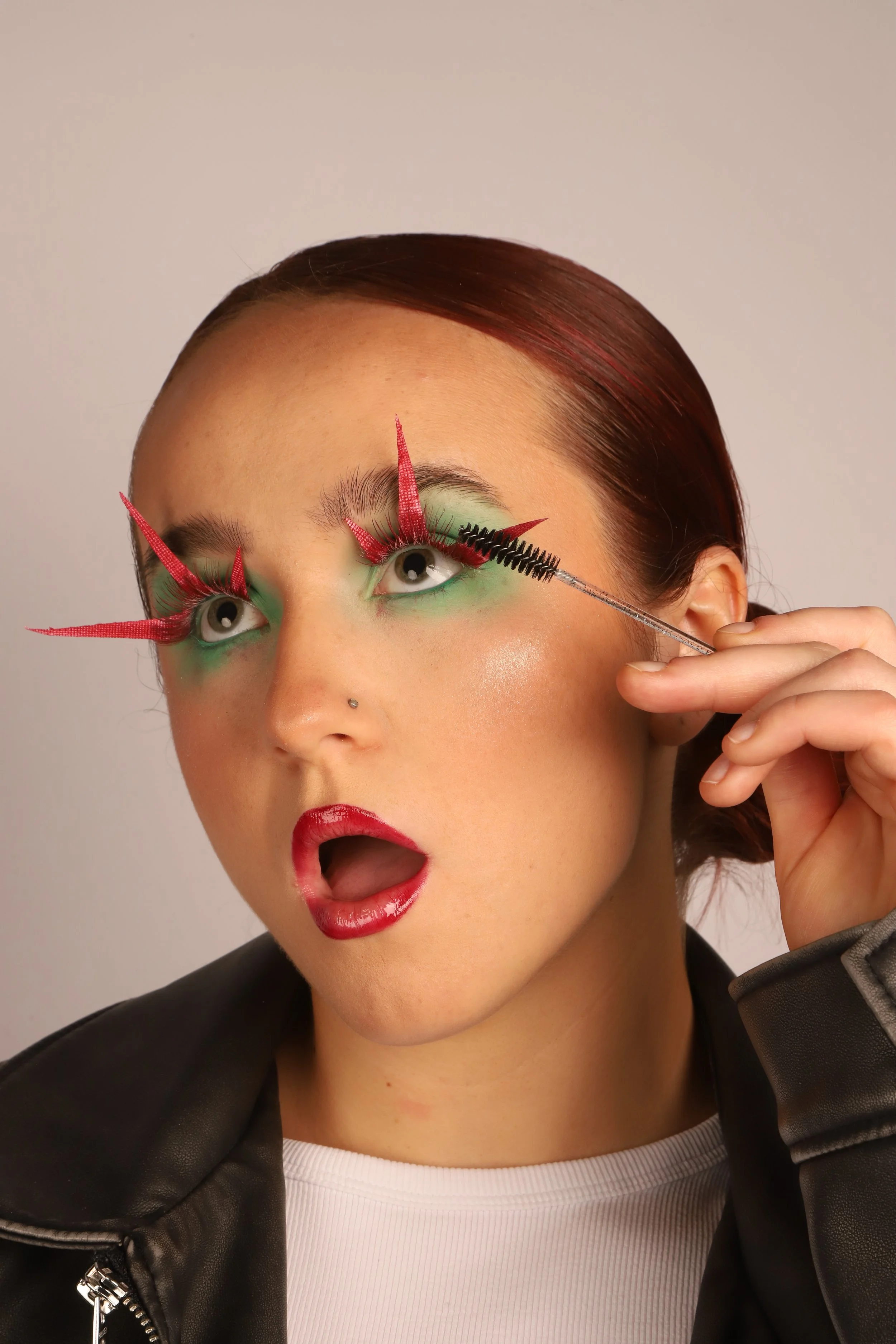 A woman with creative makeup and eyelash extensions that resemble red spikes, applying black mascara with a special wand.