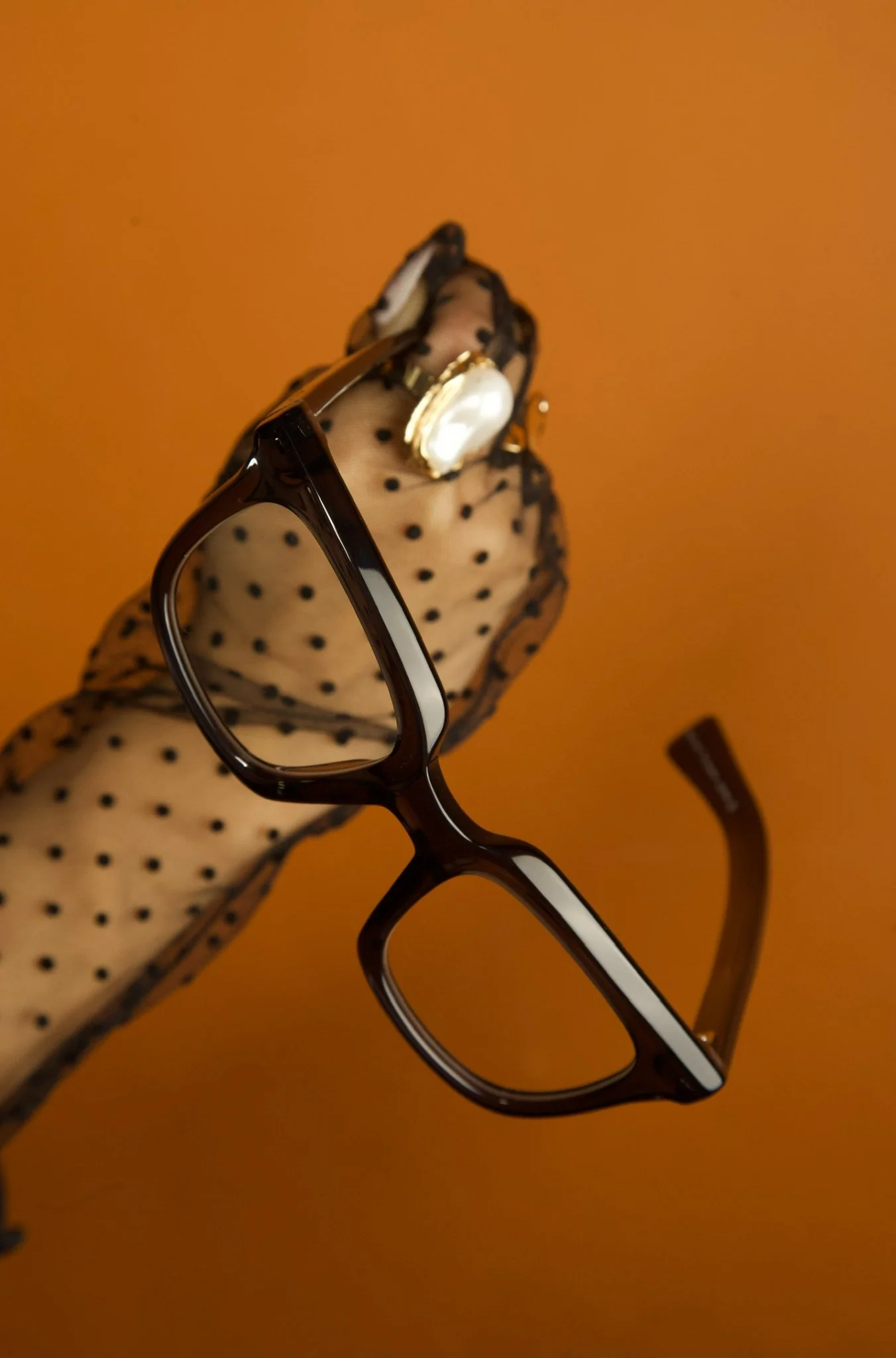 A woman wearing a sheer, polka-dotted glove holding black-framed eyeglasses and large pearl earrings against an orange background.