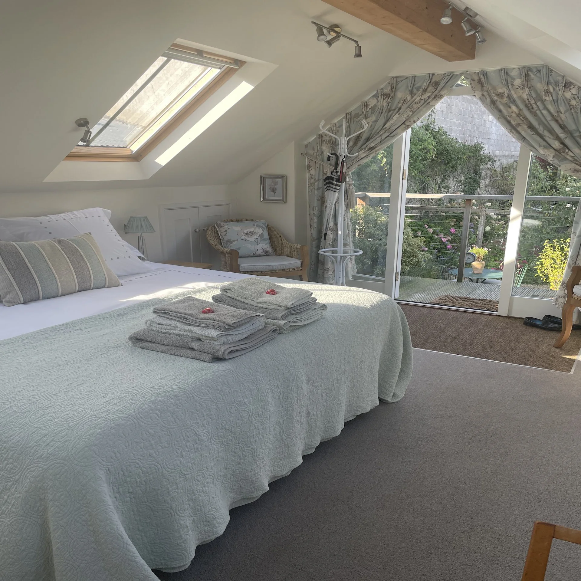 A cozy bedroom with a neatly made bed featuring white bedding and pillows, placed near a large glass door leading to a balcony with garden view, inside a room with a sloped ceiling, skylight, and wooden beam lighting fixture.