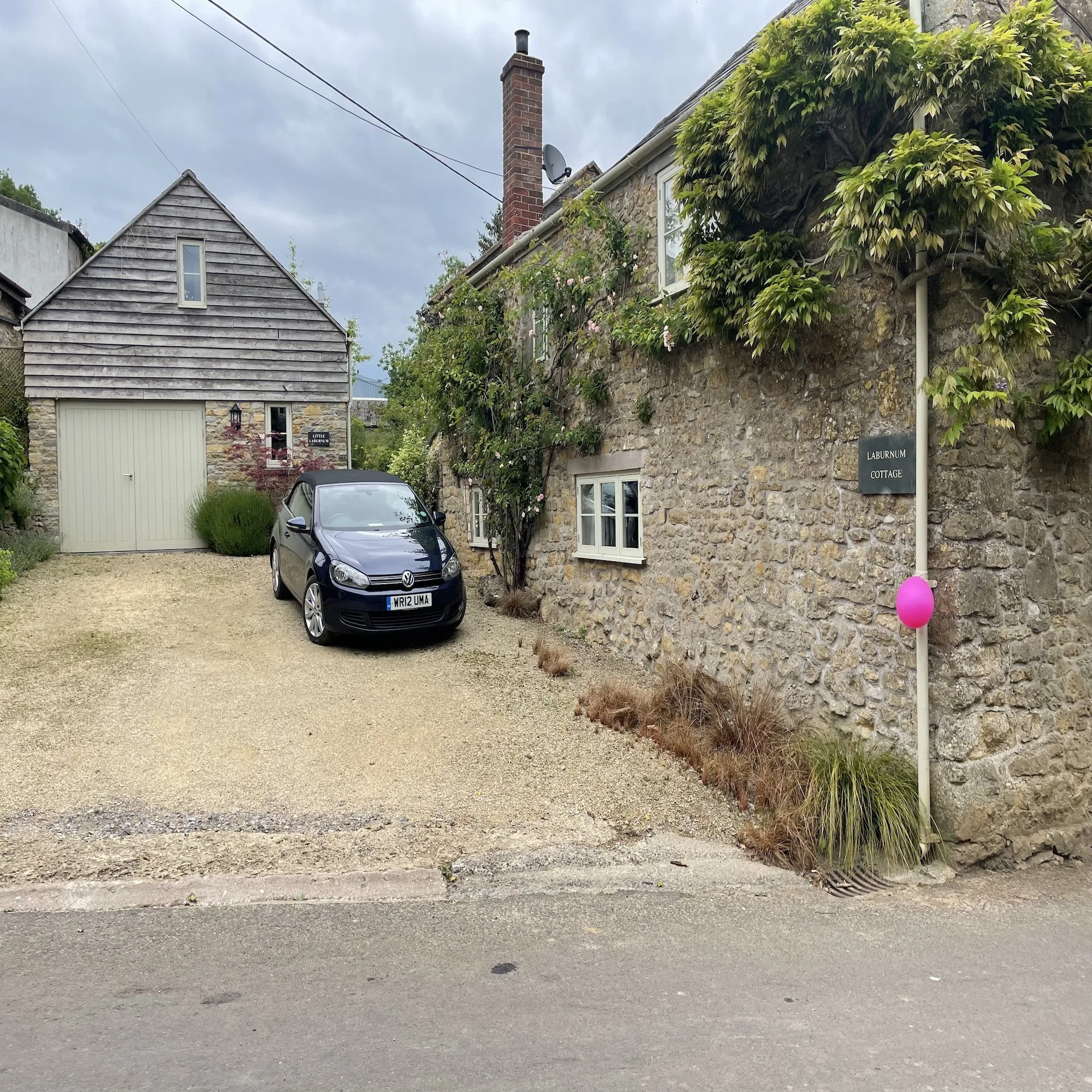 A gravel driveway in front of an old stone cottage with white window frames, decorated with climbing plants, and a small garage with wood-paneled siding. A black car is parked on the driveway. There is a sign that reads 'Laburnum Cottage' and a pink balloon tied to a pole near the cottage.