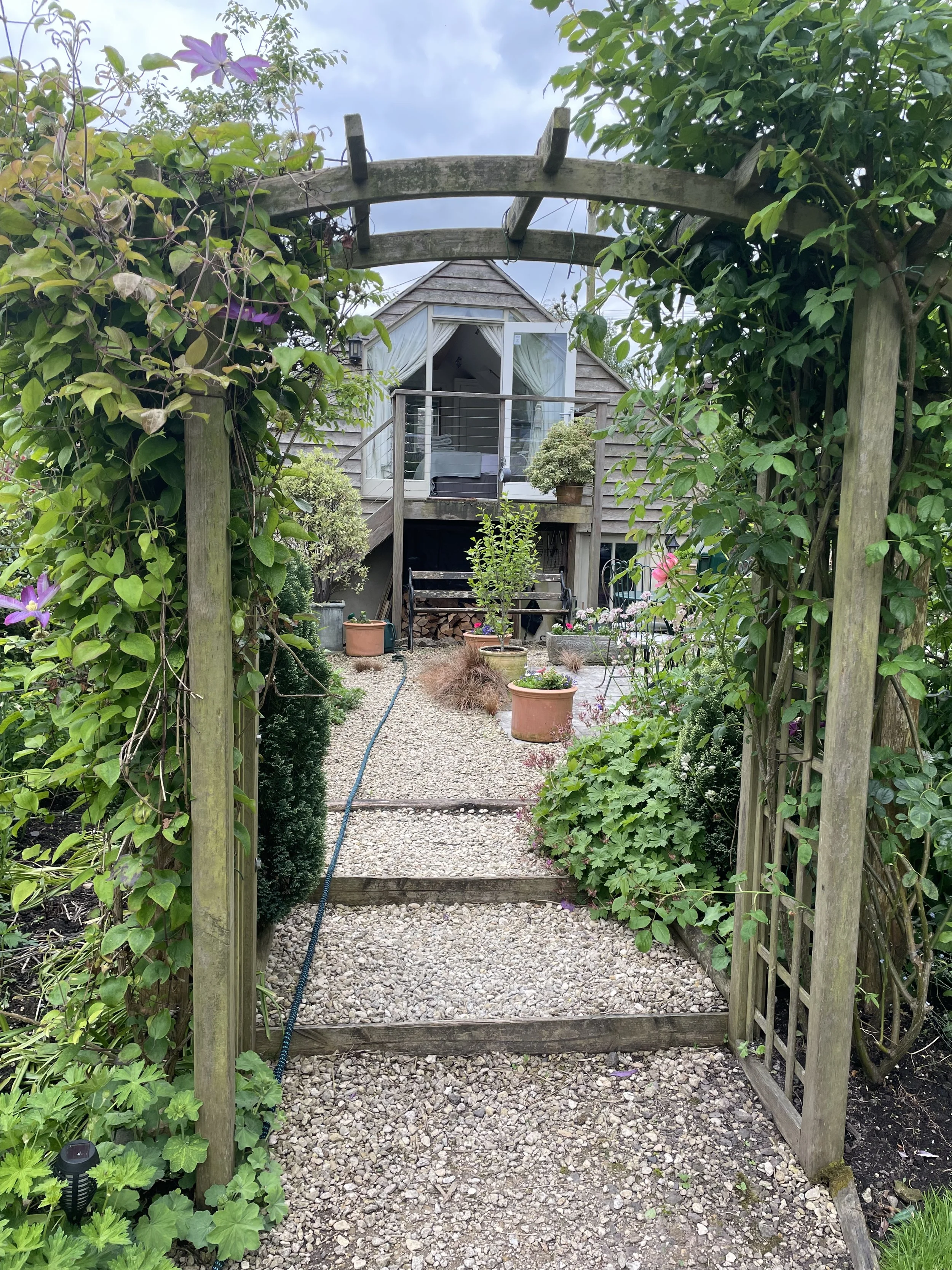 View of a garden pathway leading to a small wooden house or shed, with a wooden arbor covered in green vines and purple flowers at the entrance.