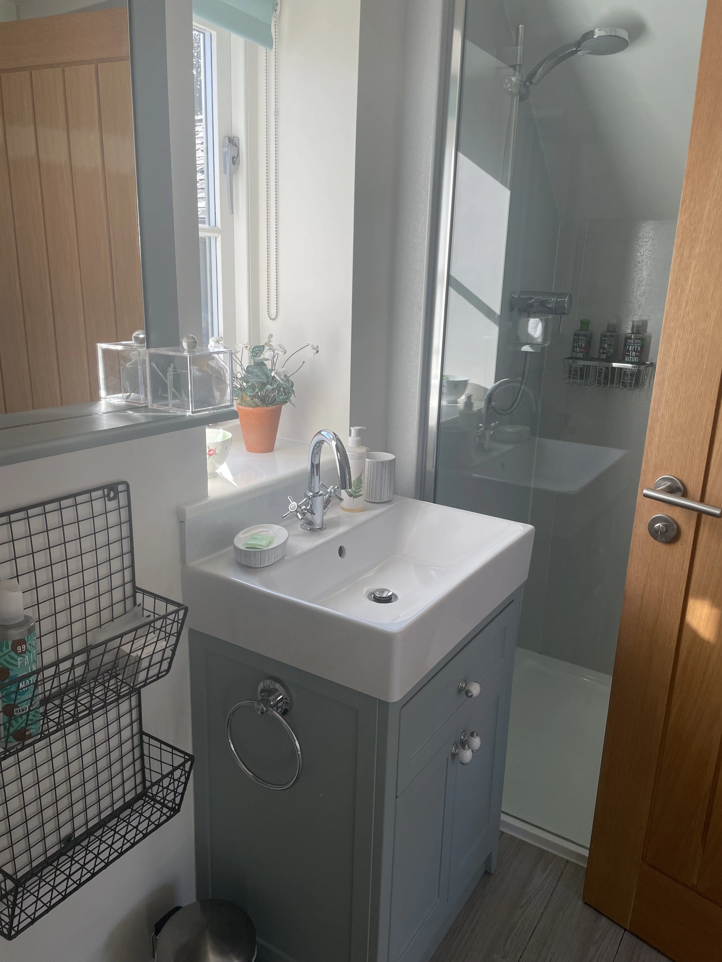 Small bathroom with a white sink, a mirror, and a window with a view outside. A small shelf holds toiletries, and a shower with a glass door is visible in the background. There is a potted plant on the window sill, and a black wire storage rack on the wall next to the sink.