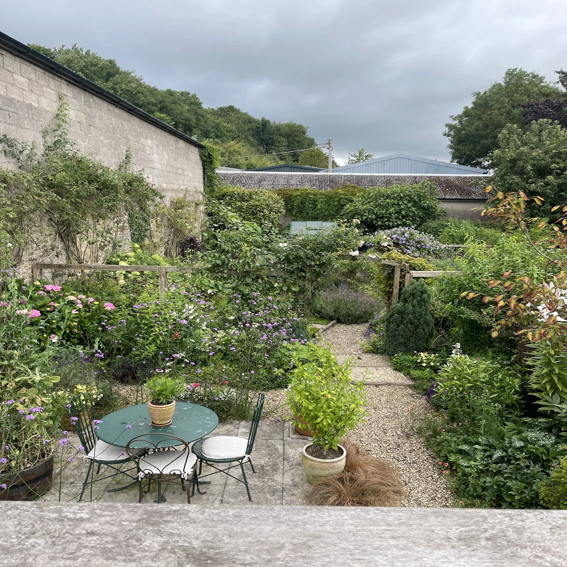 A lush garden with a variety of flowers and plants, a small gravel pathway, a patio with a green metal table and four chairs, and a stone wall in the background, under a cloudy sky.