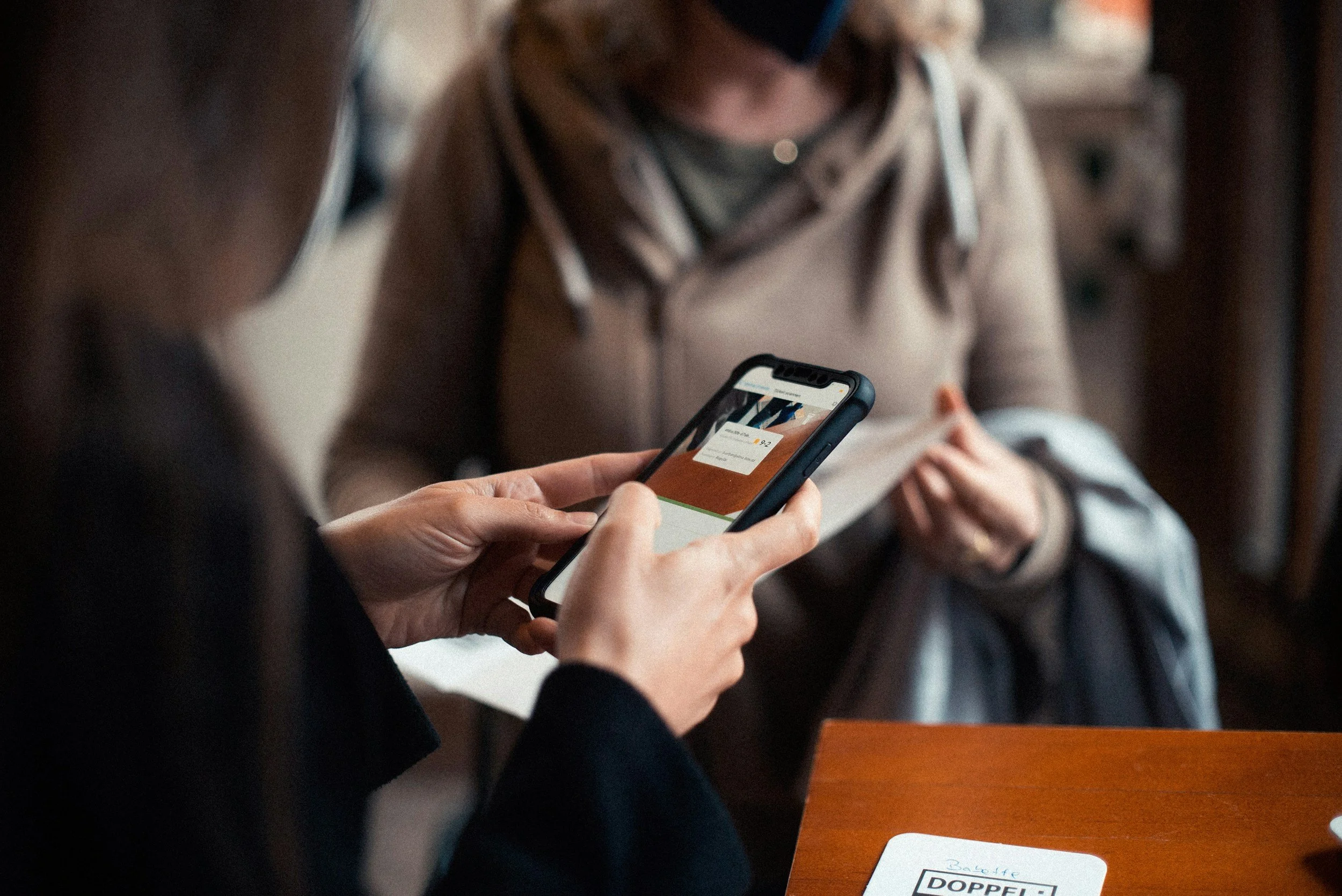 Two women sit at a table, one holding a smartphone with a physical card or ticket displayed on the screen, in an indoor setting.