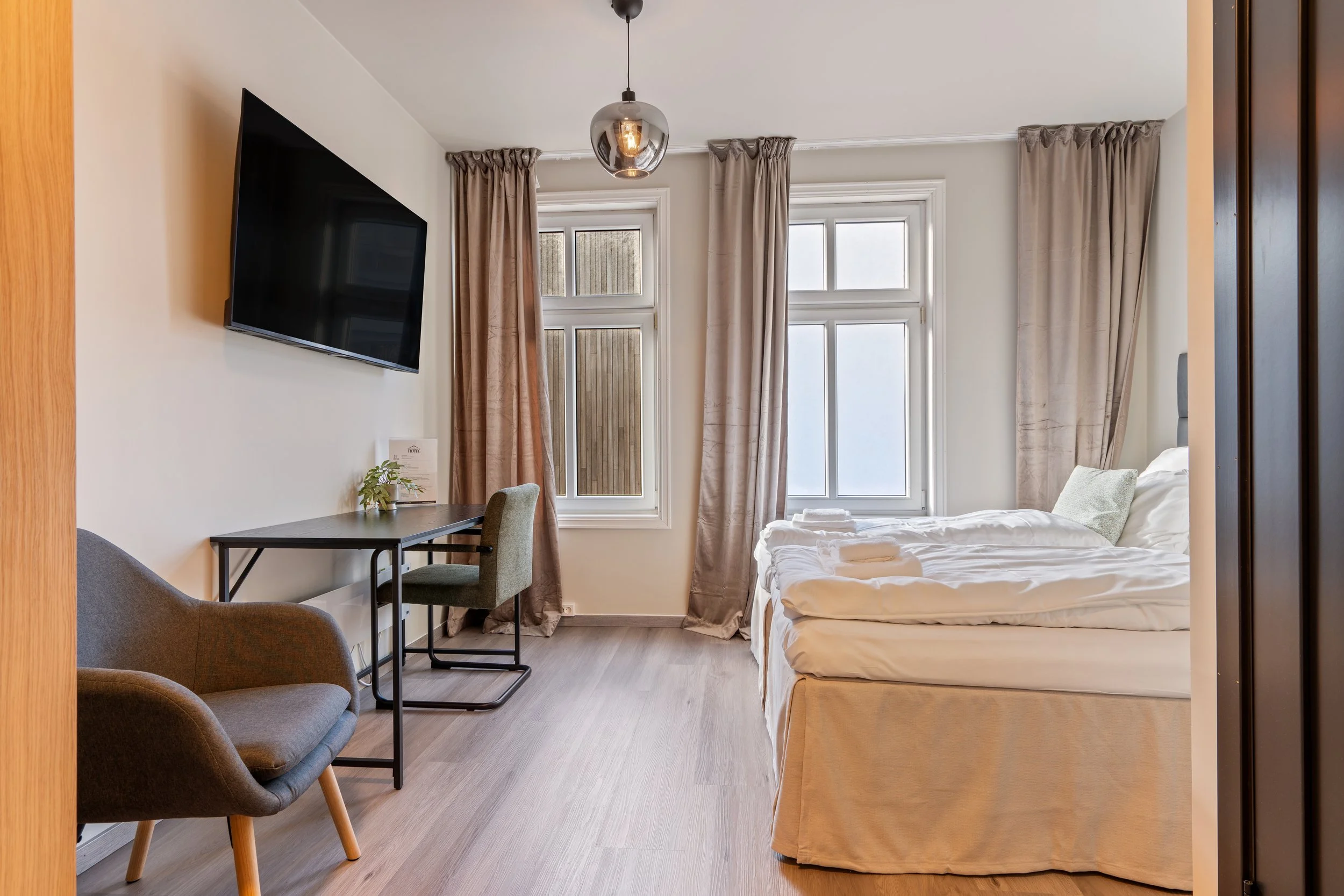 A minimalist hotel room with a bed, a wall-mounted flat-screen TV, a small desk with a chair, and large windows with beige curtains.